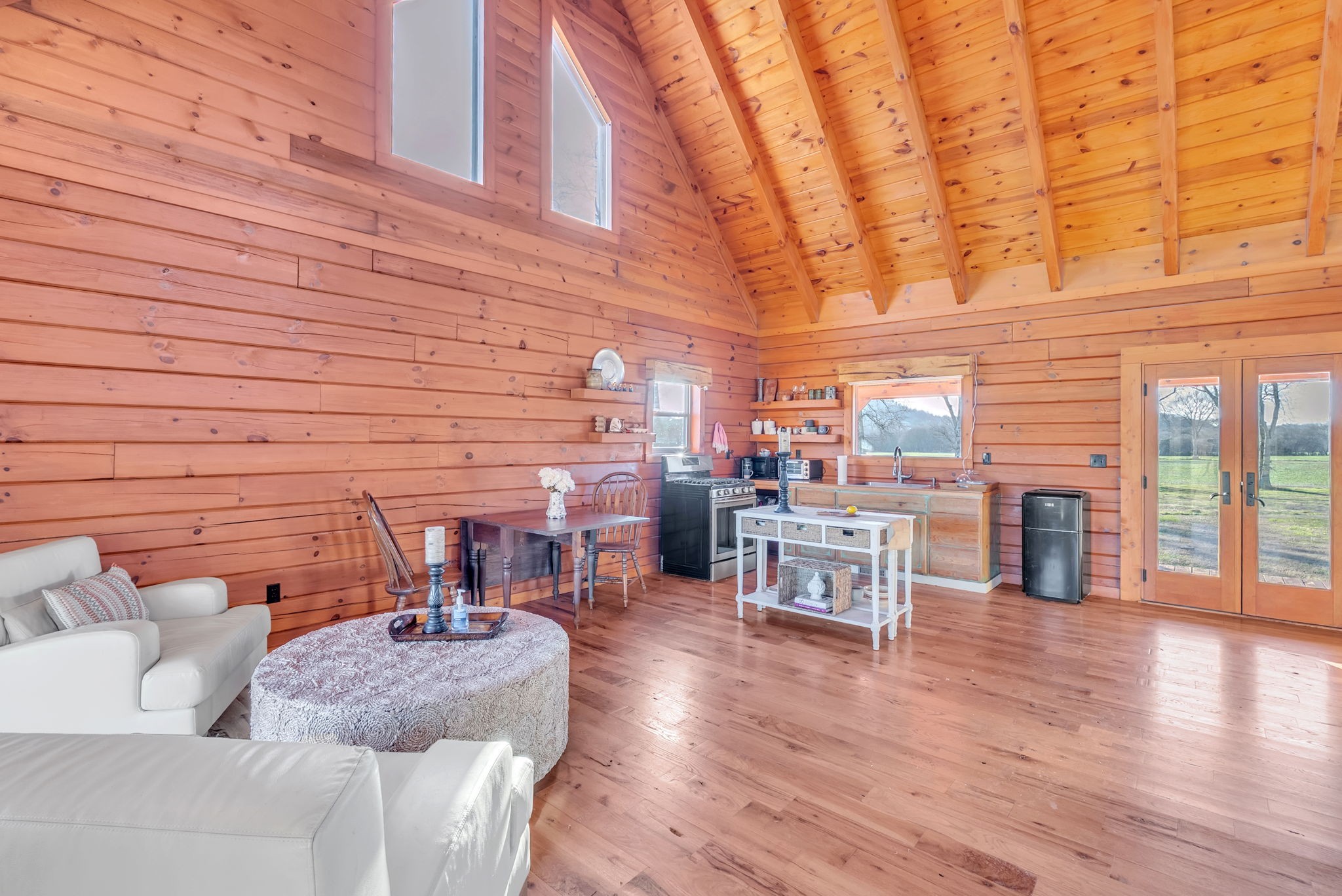 Open-plan living room and kitchen with a vaulted wood ceiling, hardwood floors, and glass doors to the lawn.