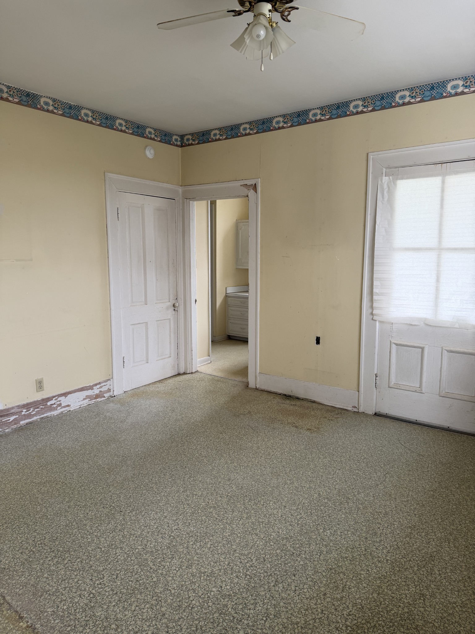 Empty room with patterned wallpaper border, worn trim, speckled floor, ceiling fan, and window blinds.