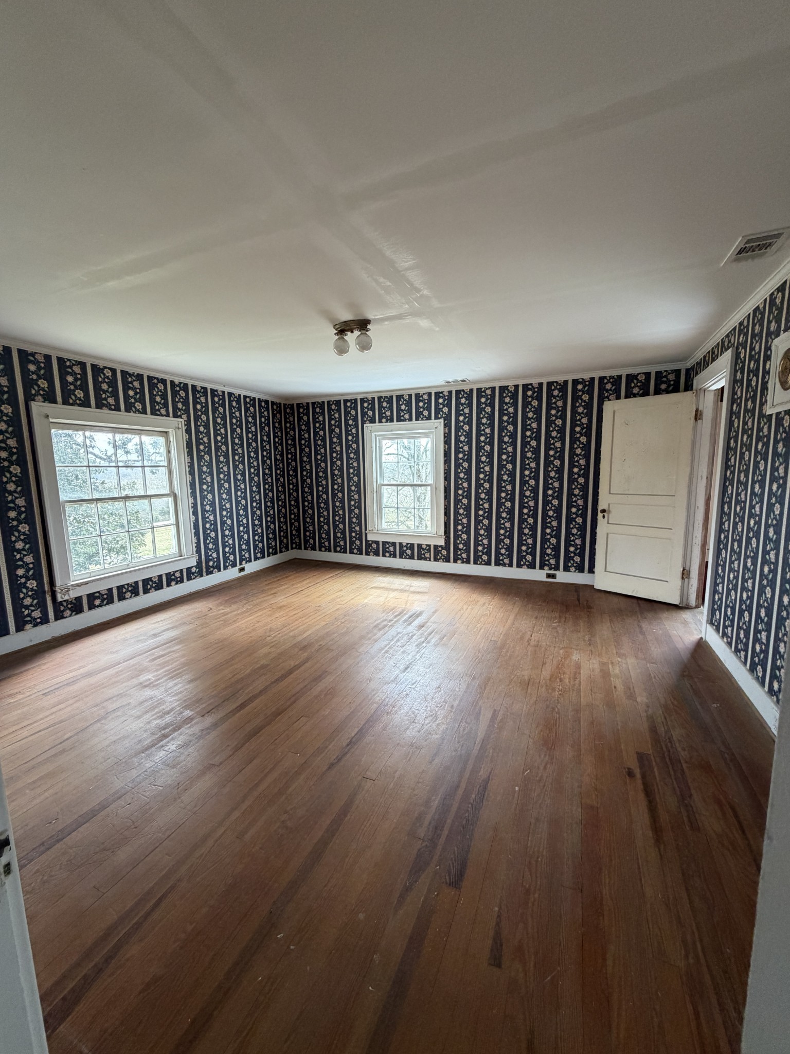Empty room with dark floral striped wallpaper, two windows, and worn hardwood floors.