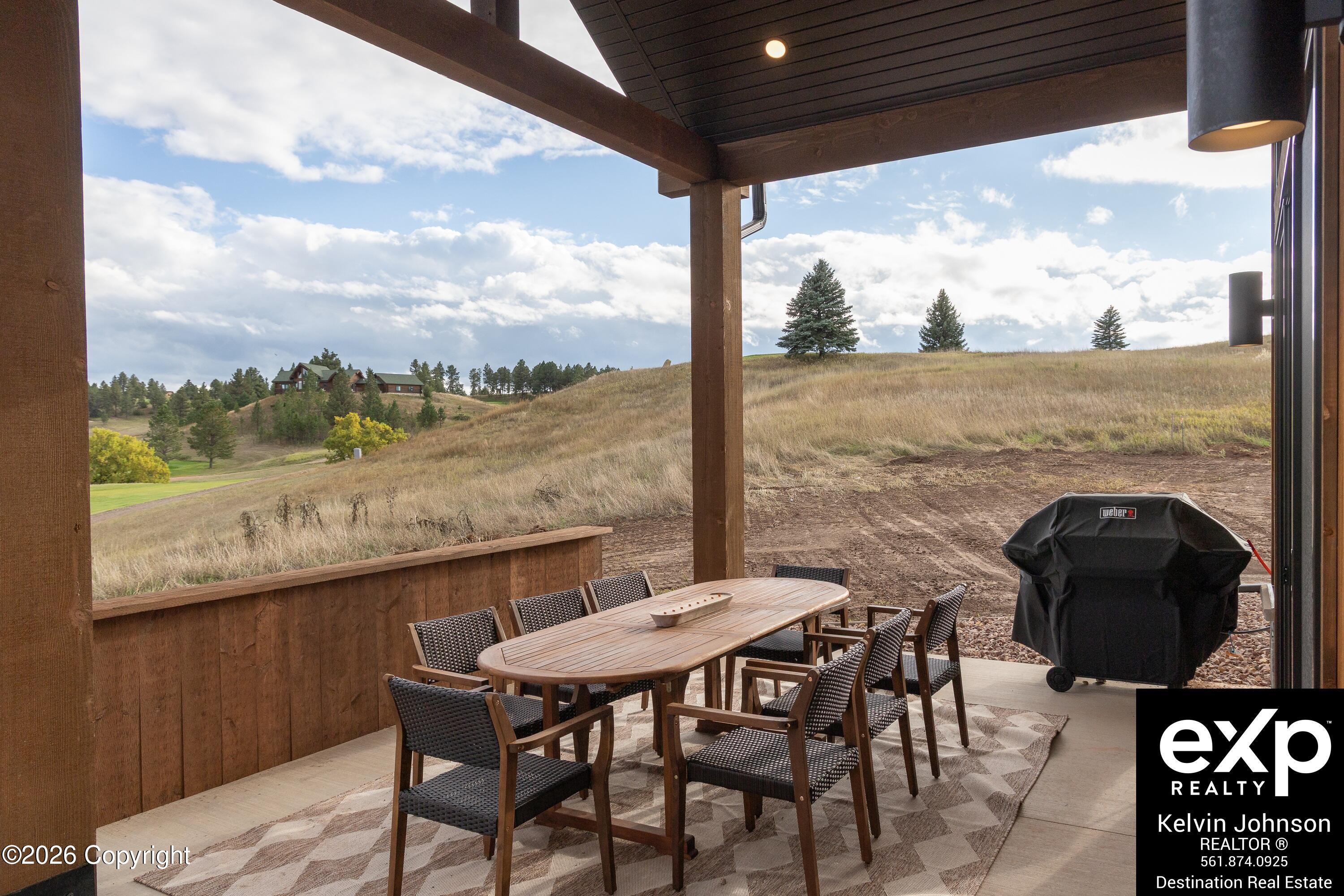 Covered patio with outdoor dining set and grill overlooking a grassy hillside and distant home.