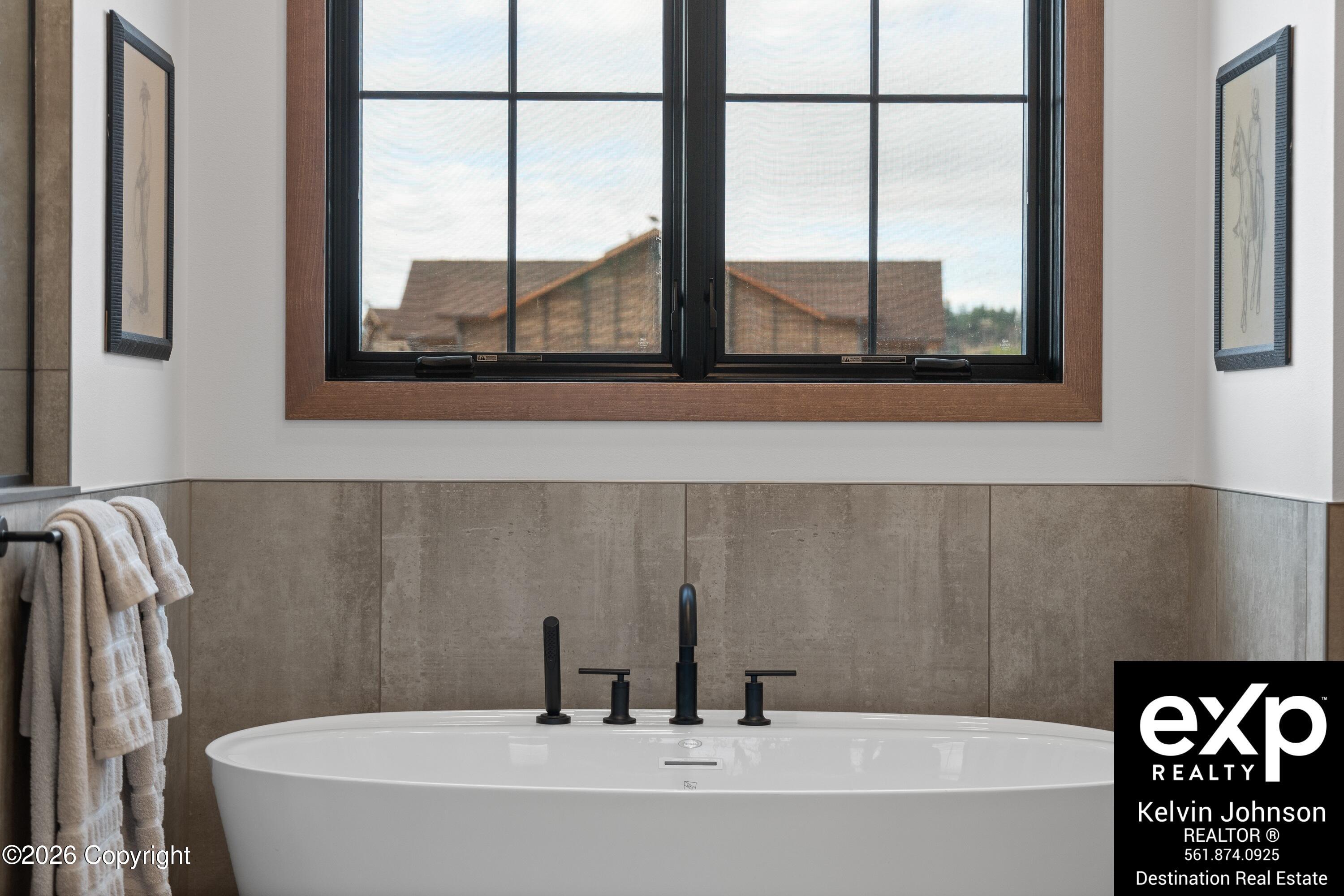 Freestanding white bathtub beneath a large window, with taupe tile and matte-black fixtures.