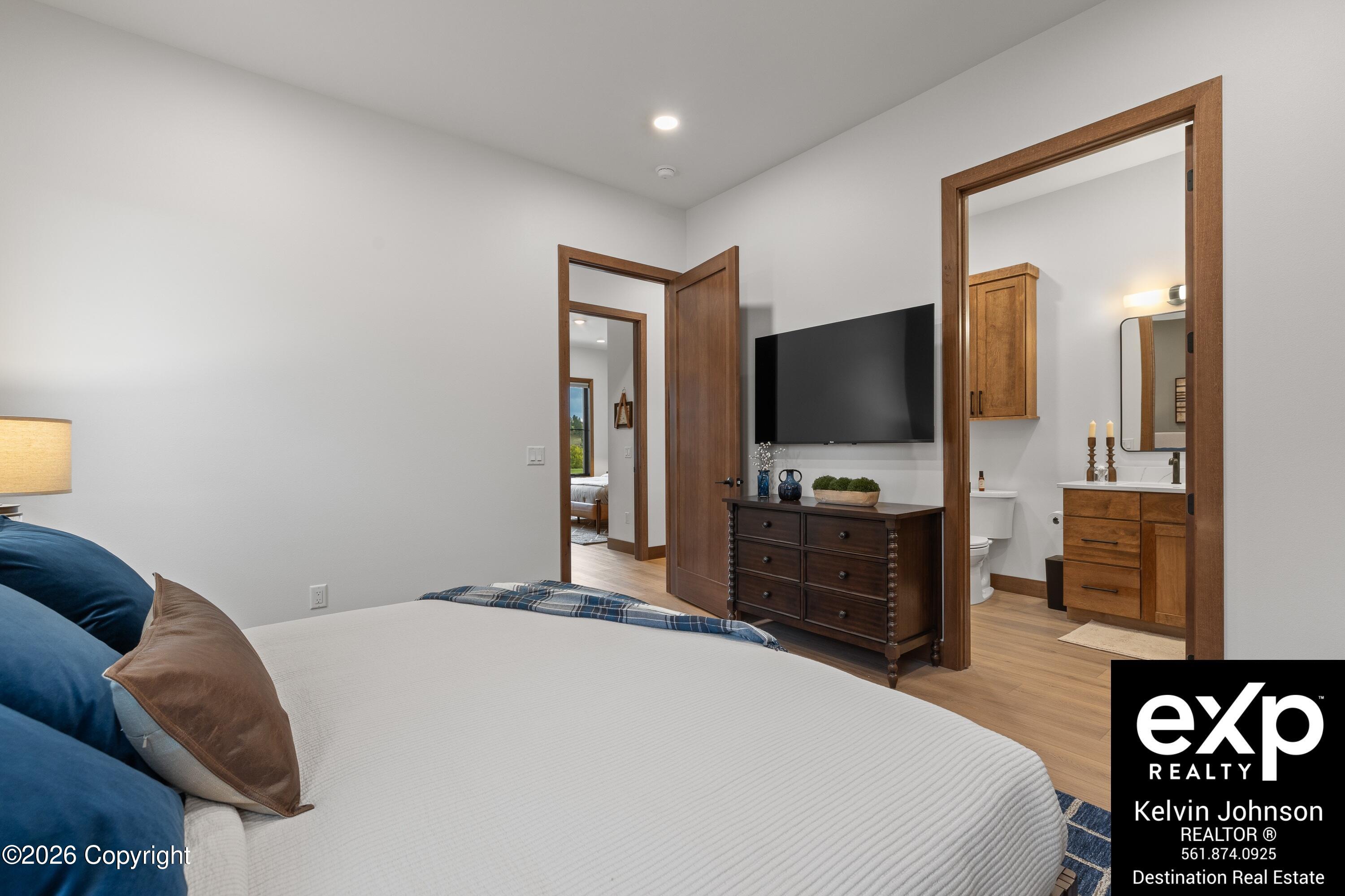 Furnished bedroom with a wall-mounted TV, dark dresser, and open doorway to an en-suite bathroom.