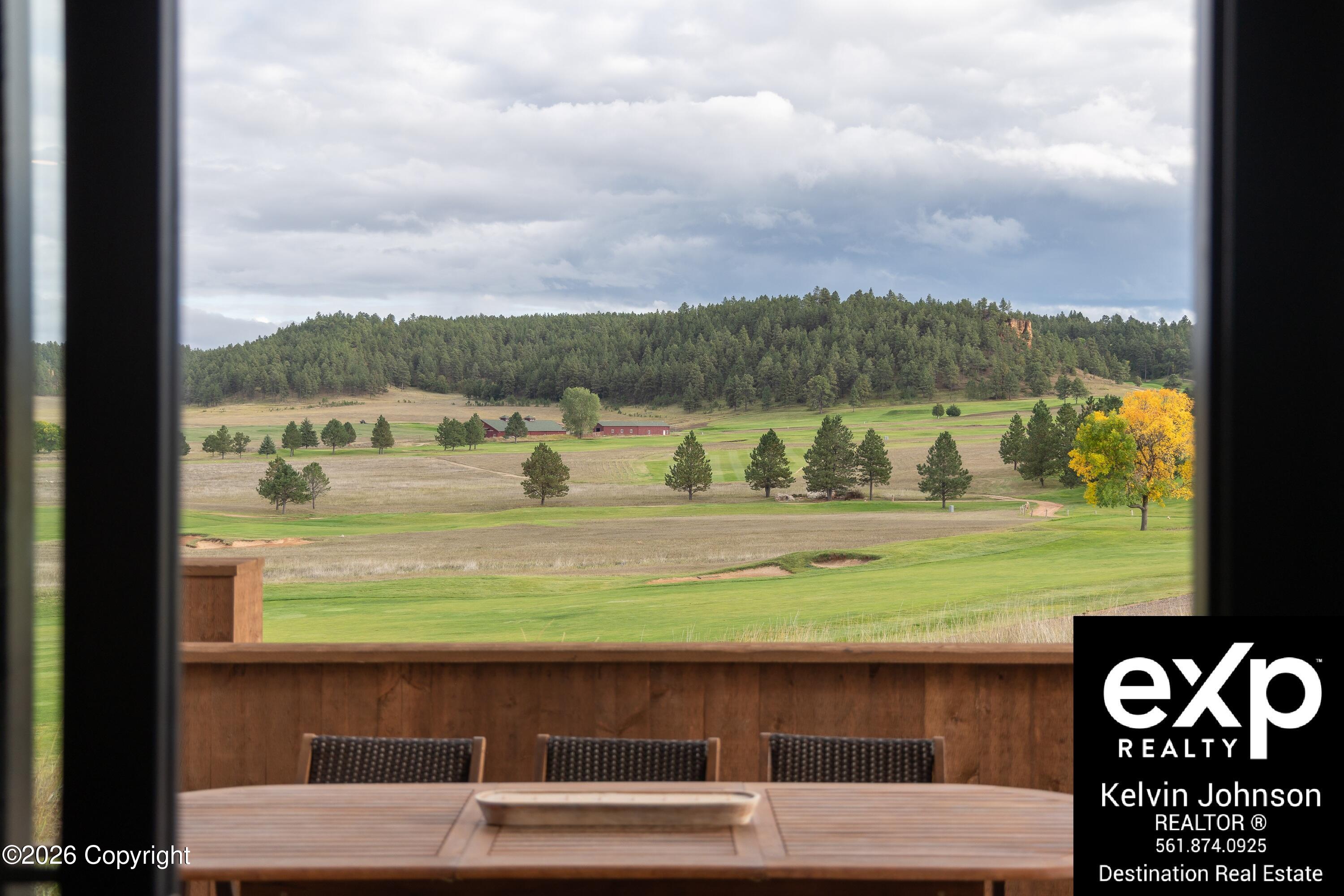 Golf course view from a deck, with a wooden dining set, red farm buildings, and a forested ridge.