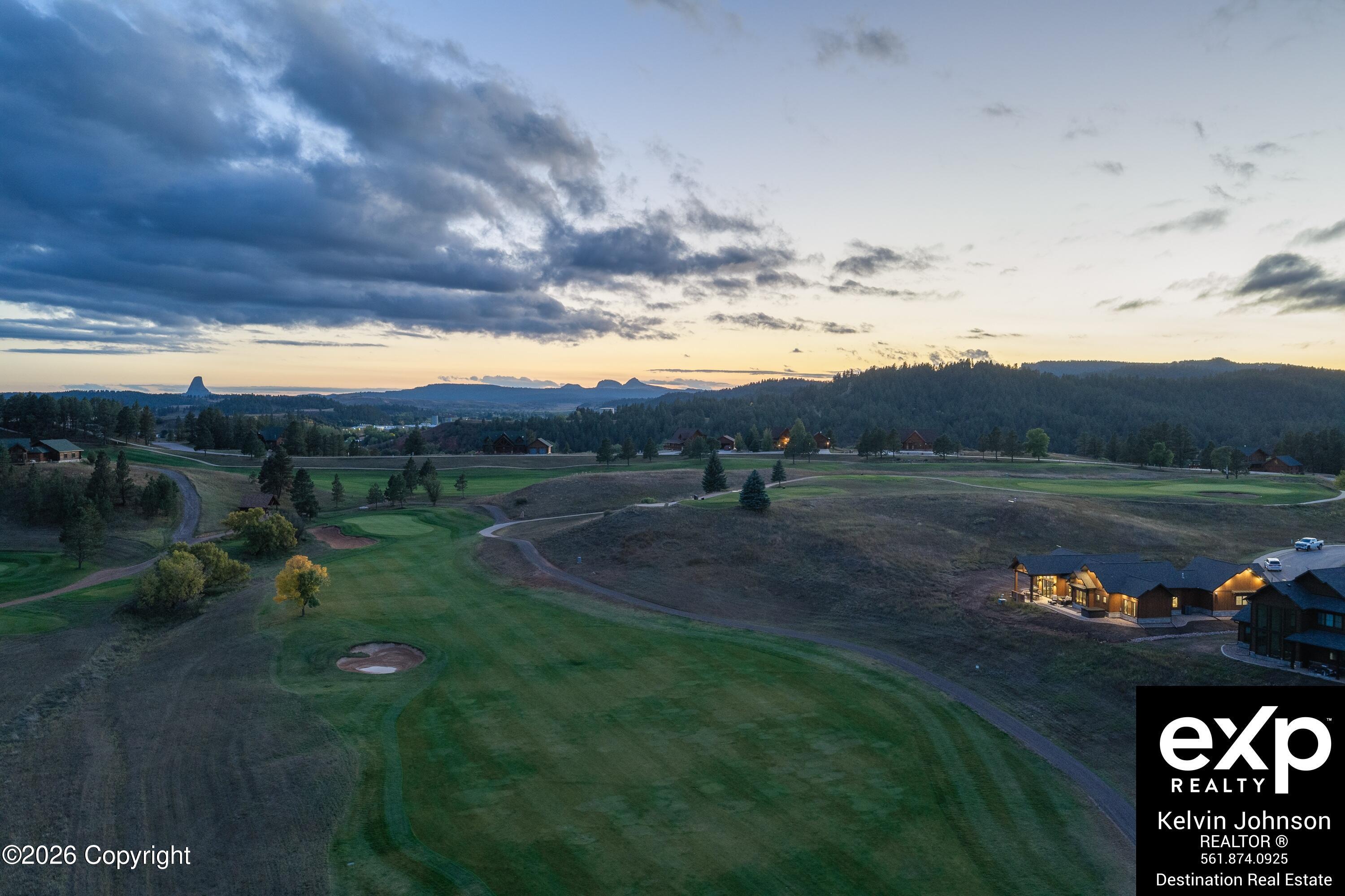 Golf course at dusk with a lit building, wooded hills, and a distinctive rock formation on the horizon.