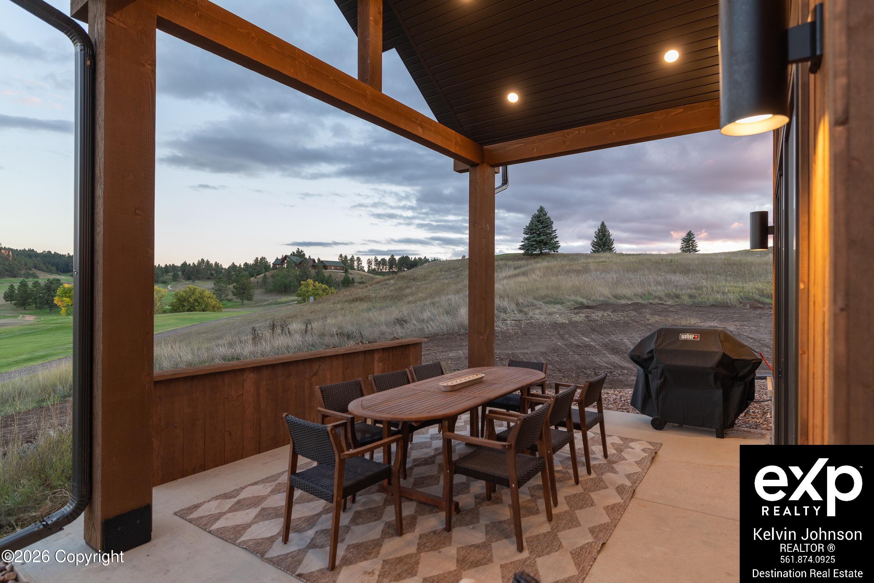 Covered patio with outdoor dining set and grill overlooking grassy hills and a golf course at dusk.