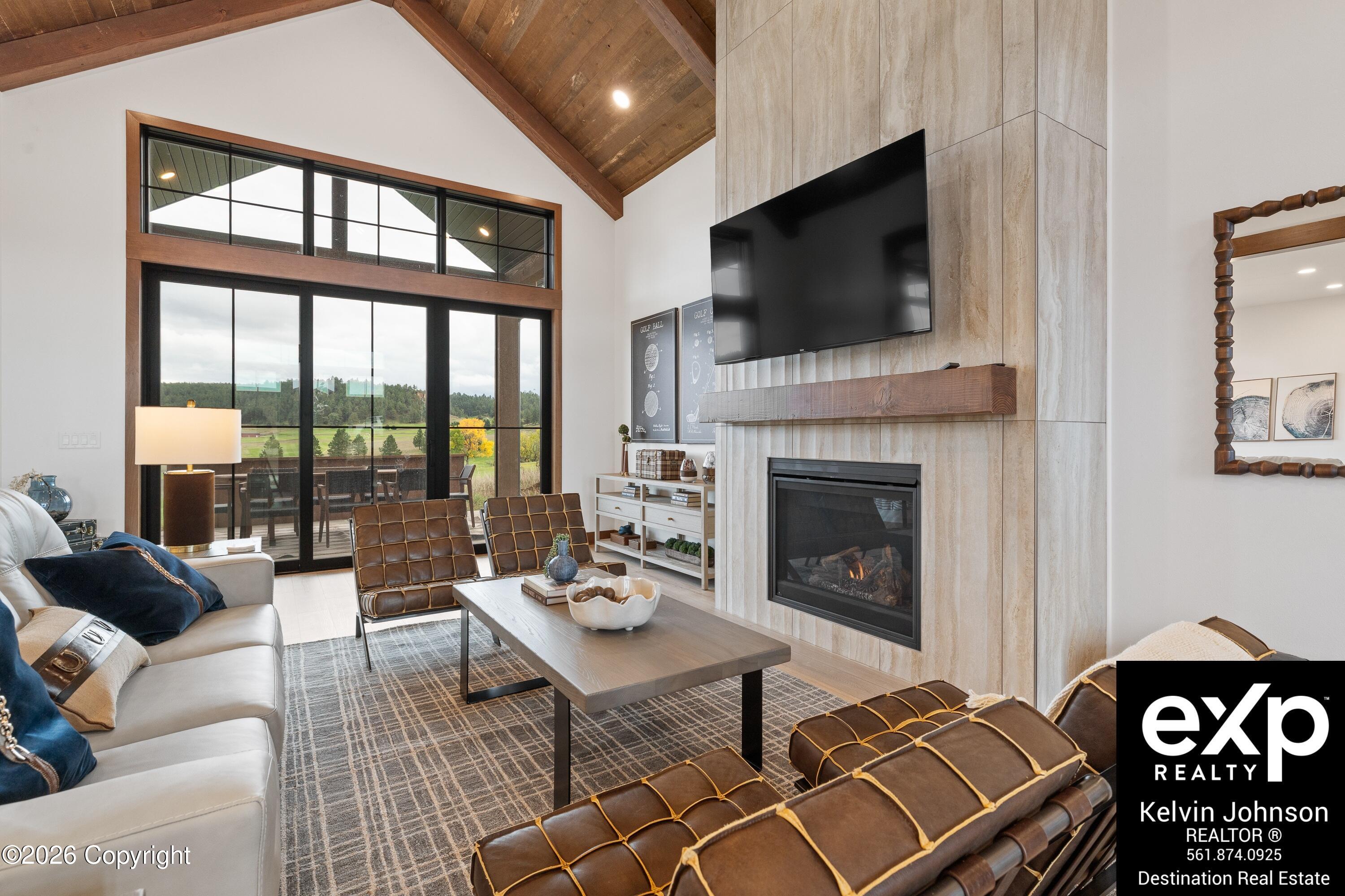 Living room with vaulted wood ceiling, stone fireplace, leather chairs, and tall windows overlooking a deck.