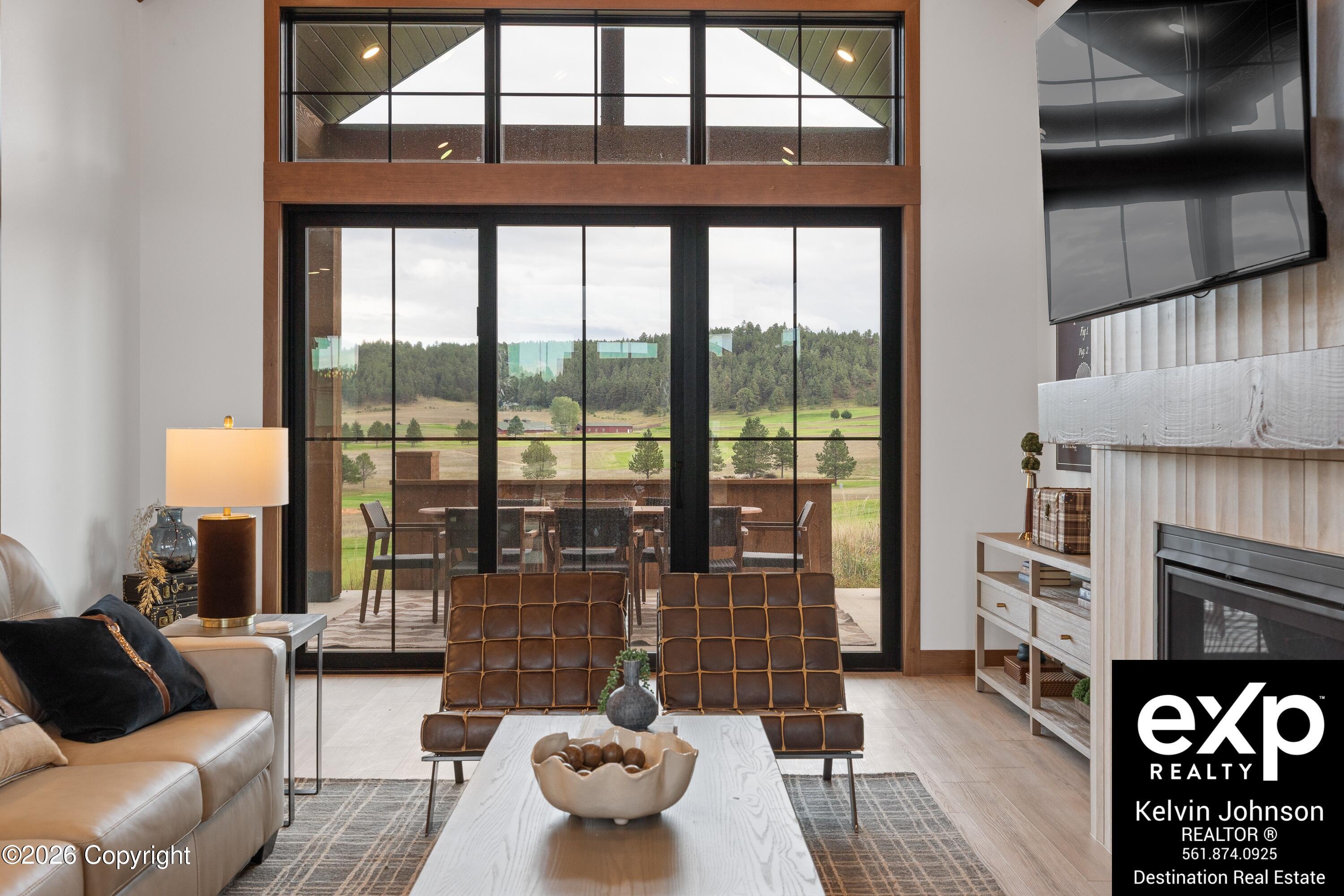 Furnished living room with tall glass doors overlooking a patio, open fields, and a wooded hillside.