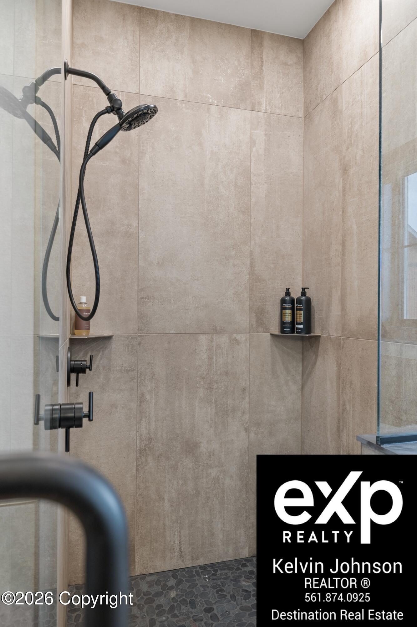 Walk-in shower with beige tile, matte black fixtures, glass enclosure, and dark pebble flooring.