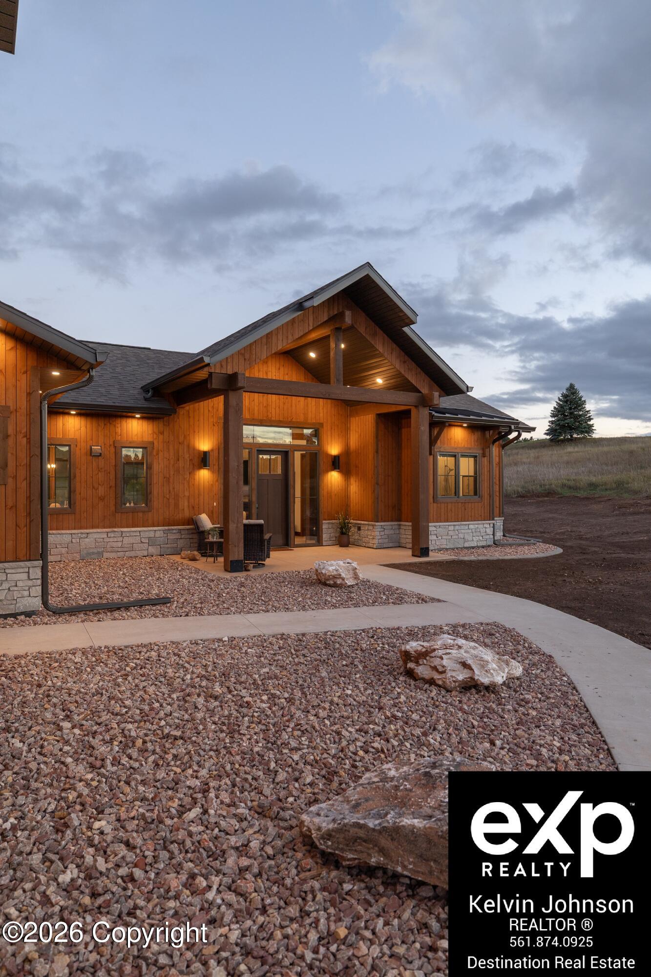 Modern wood-sided home with a warmly lit covered entrance, stone base, and rock landscaping at dusk.