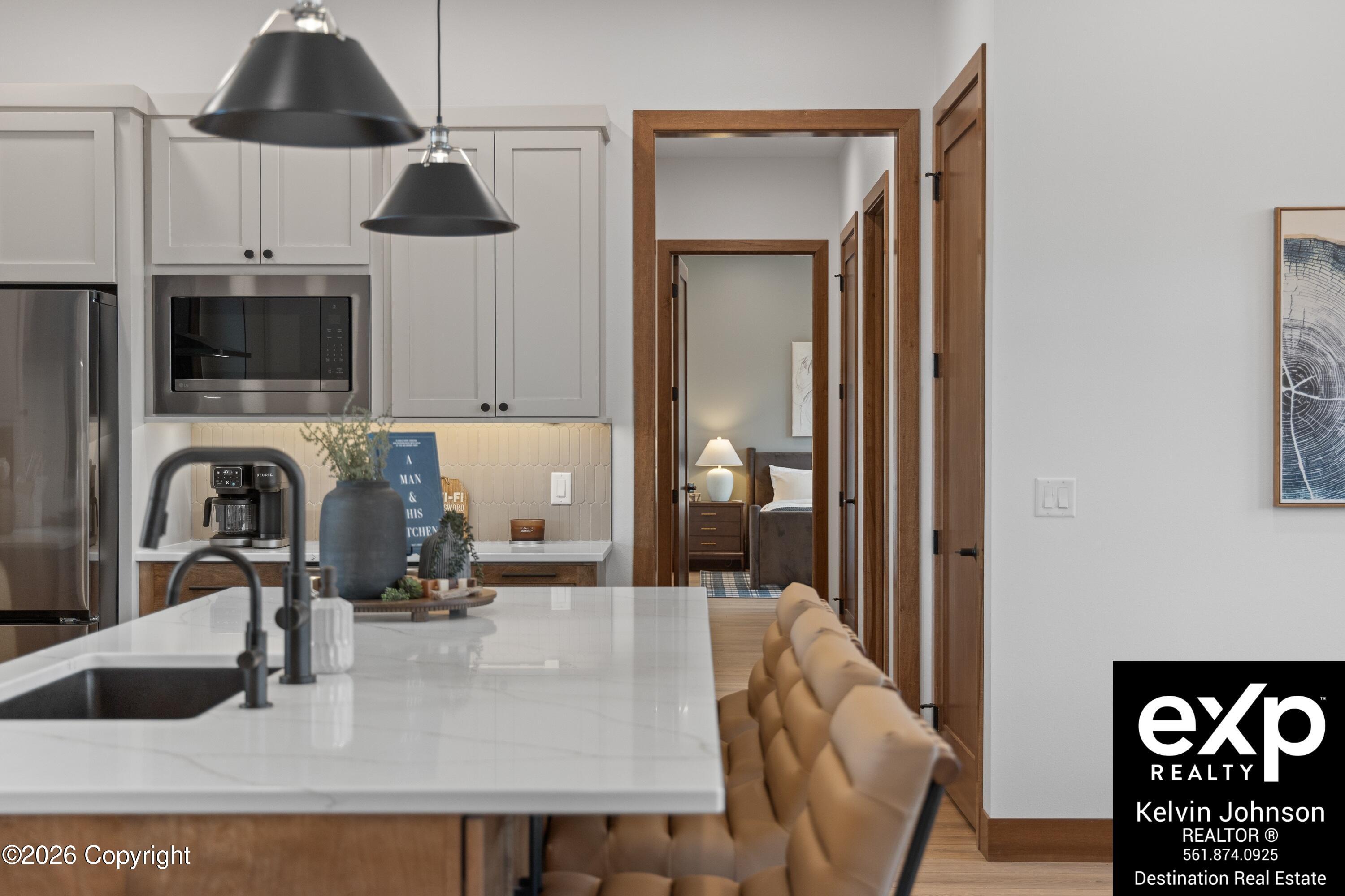 Modern kitchen with a white island, black pendant lights, and a bedroom visible through the doorway.