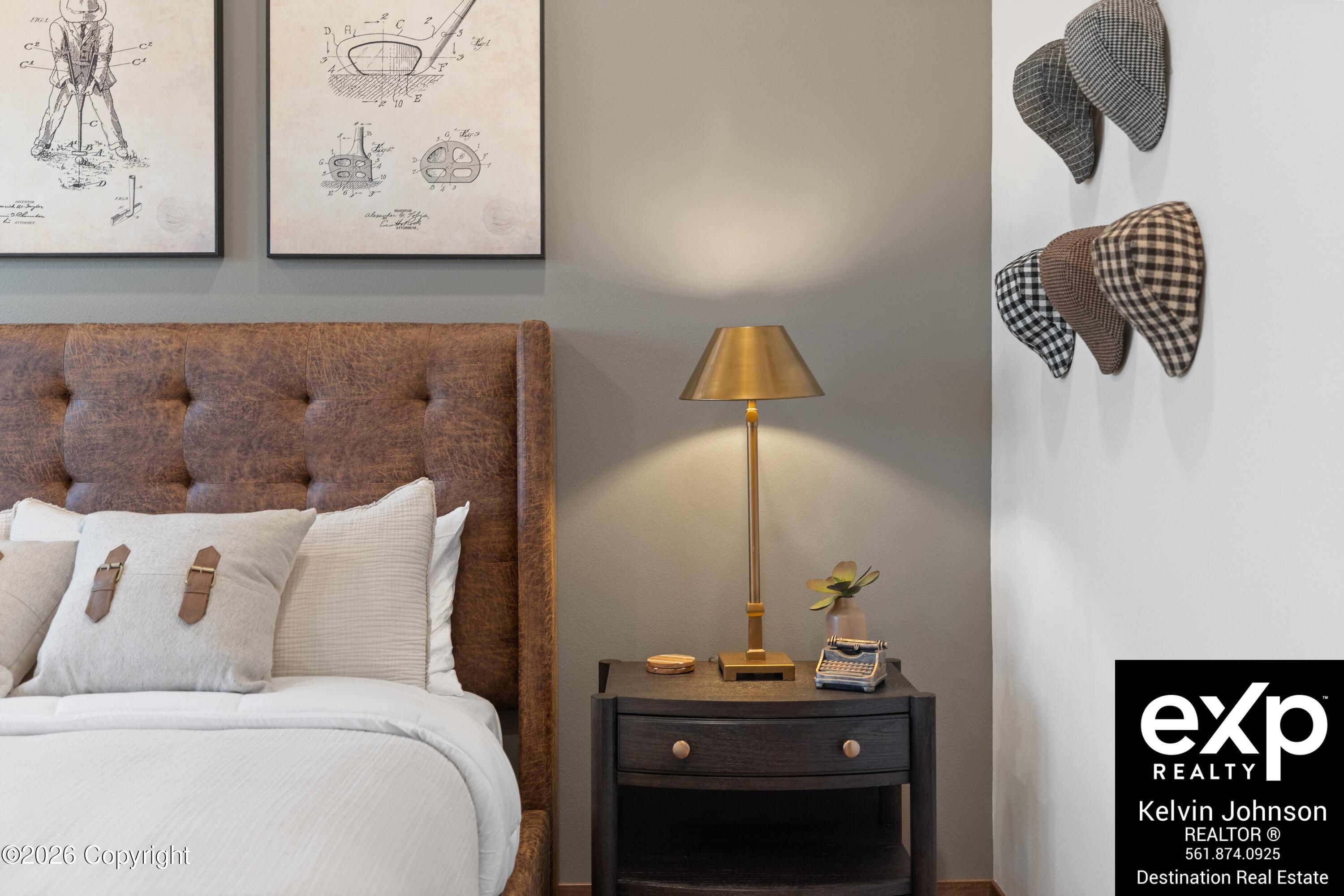 Bedroom with a brown leather headboard, brass bedside lamp, framed prints, and patterned hats on the wall.