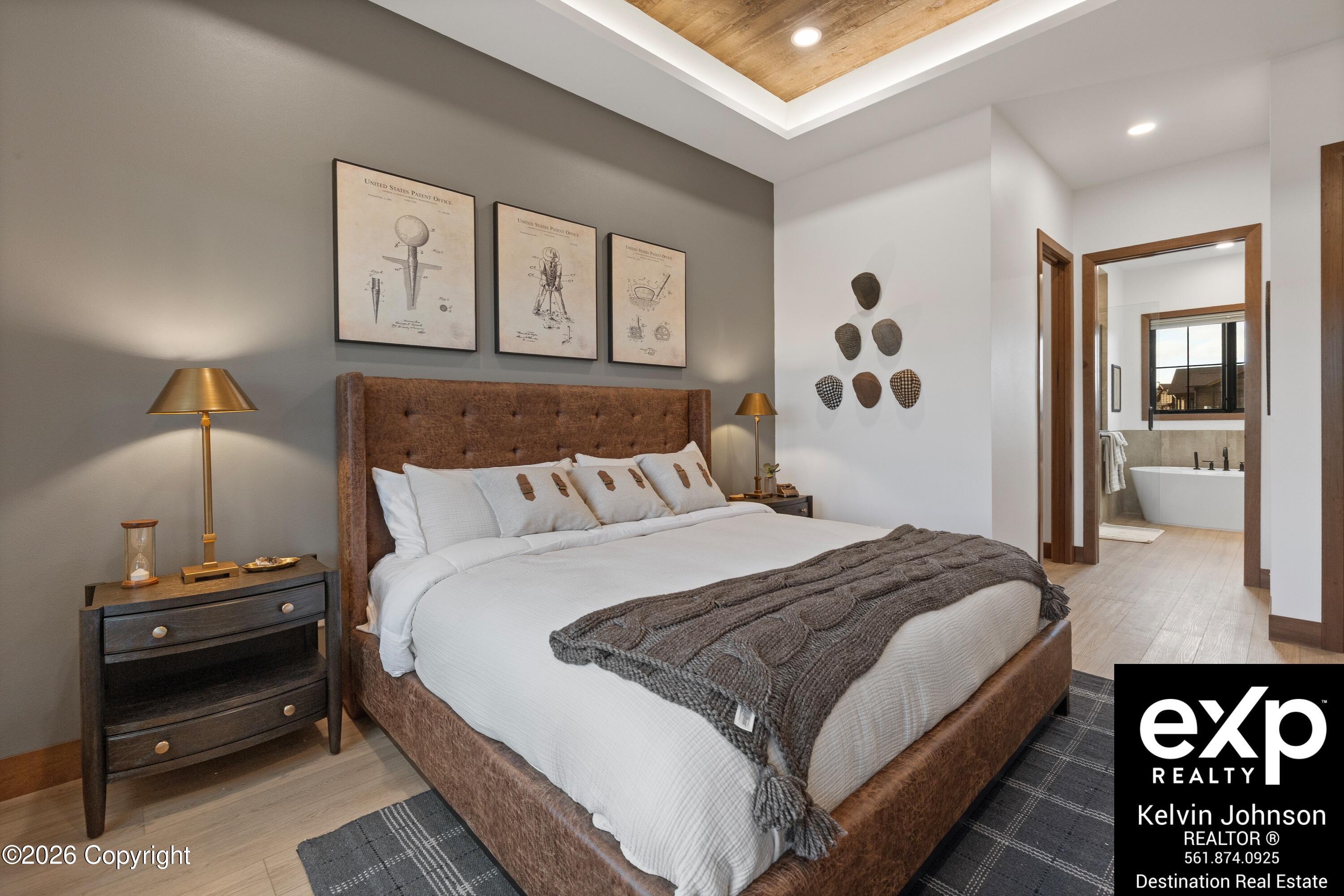 Furnished bedroom with a brown upholstered bed, gray throw, framed wall art, and ensuite bathroom visible through the doorway.