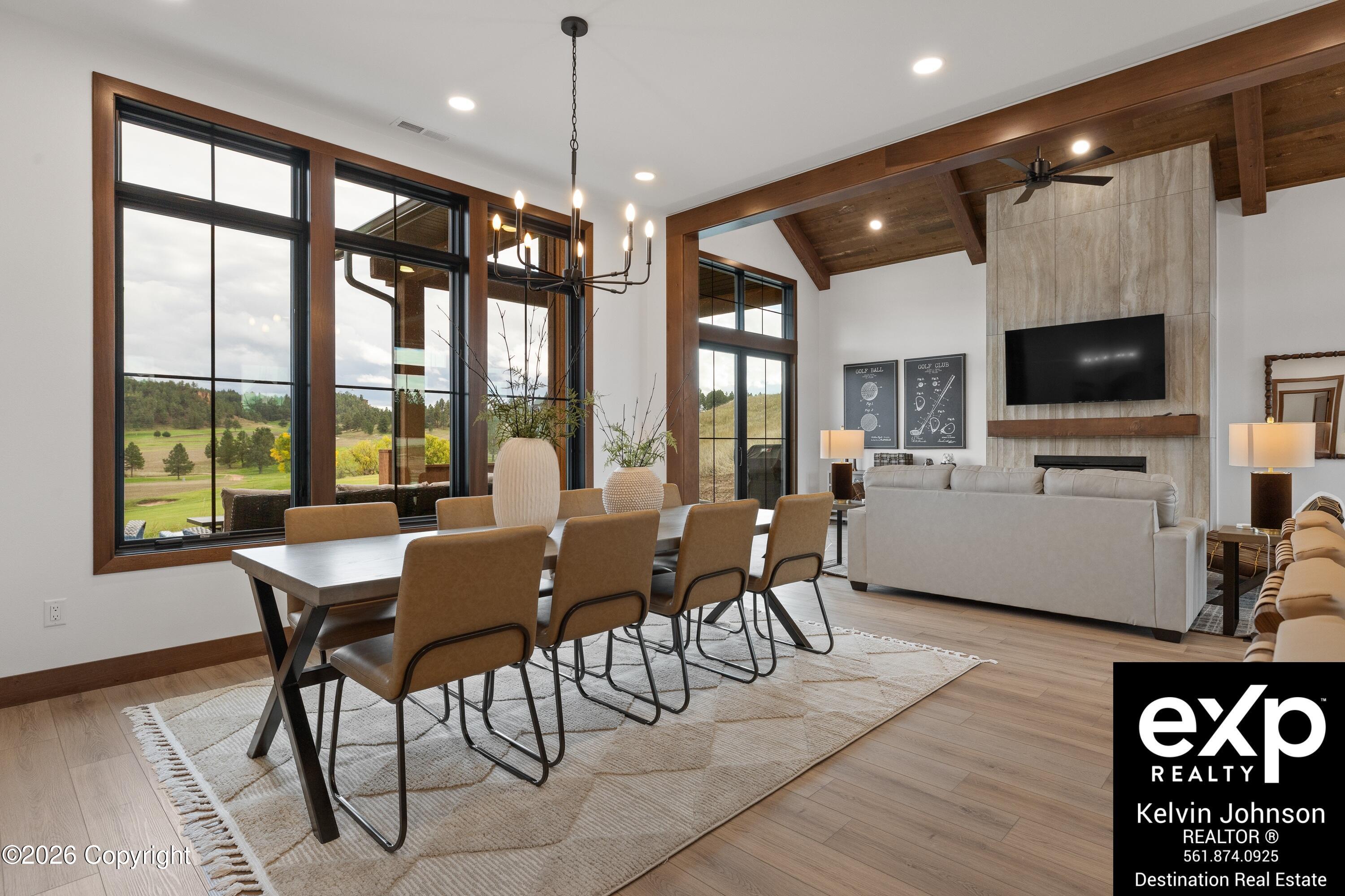 Open-plan living and dining room with large windows, a stone fireplace, and exposed wood ceiling beams.