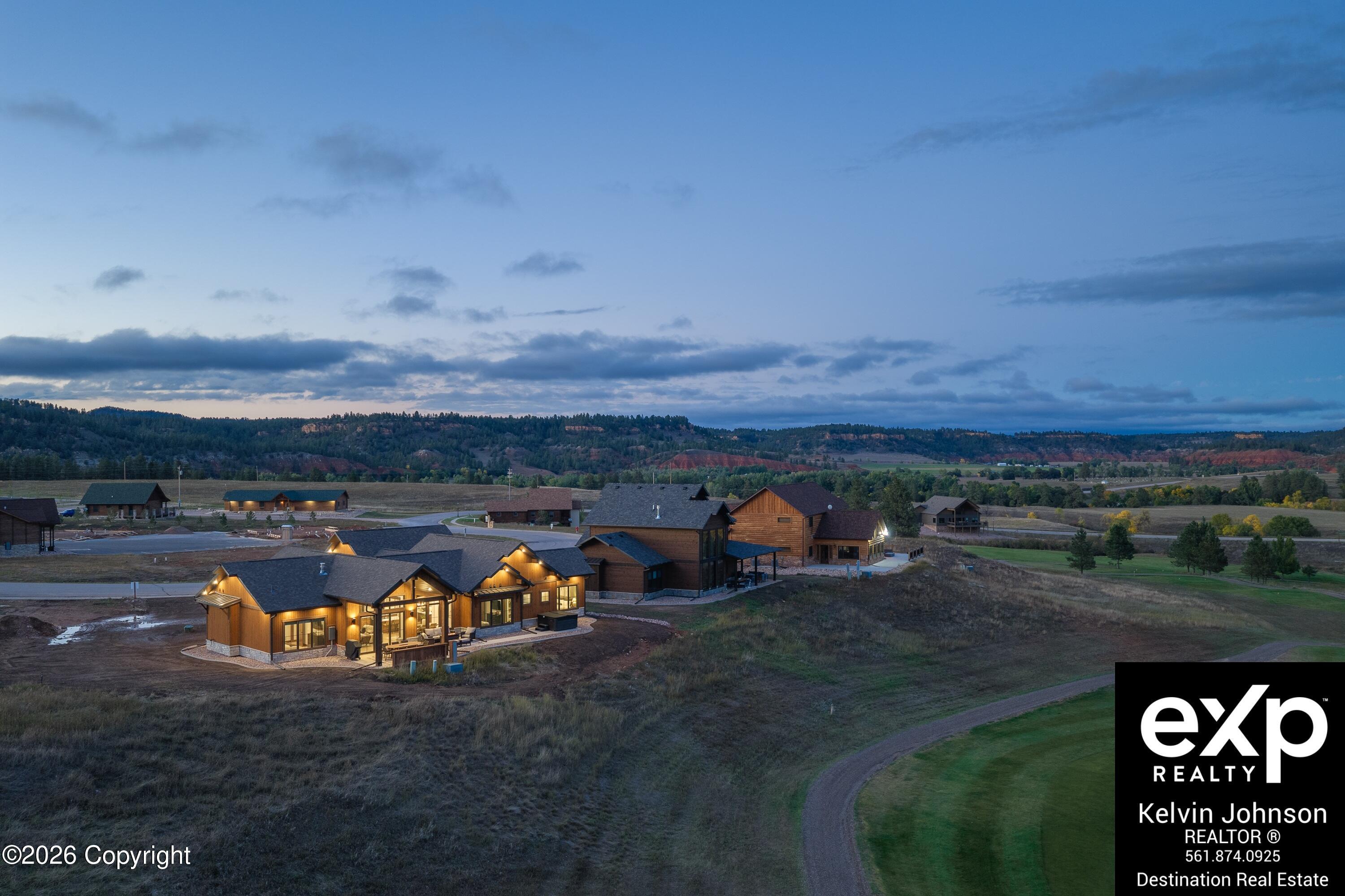 Aerial view of a warmly lit wood-sided home and neighboring houses amid open grassland and wooded hills at dusk.
