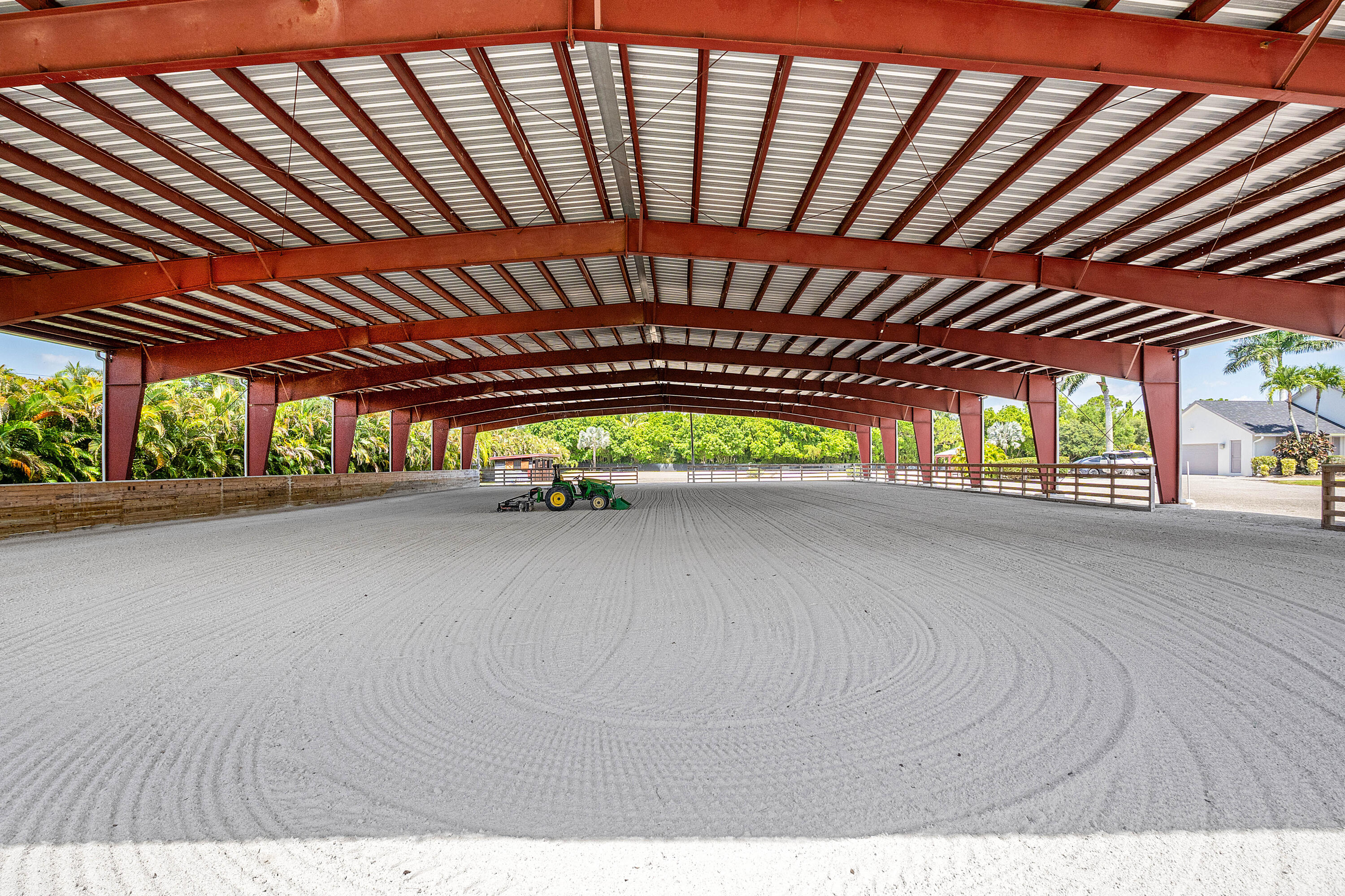 Covered riding arena with red steel beams, groomed sand, and a green tractor.