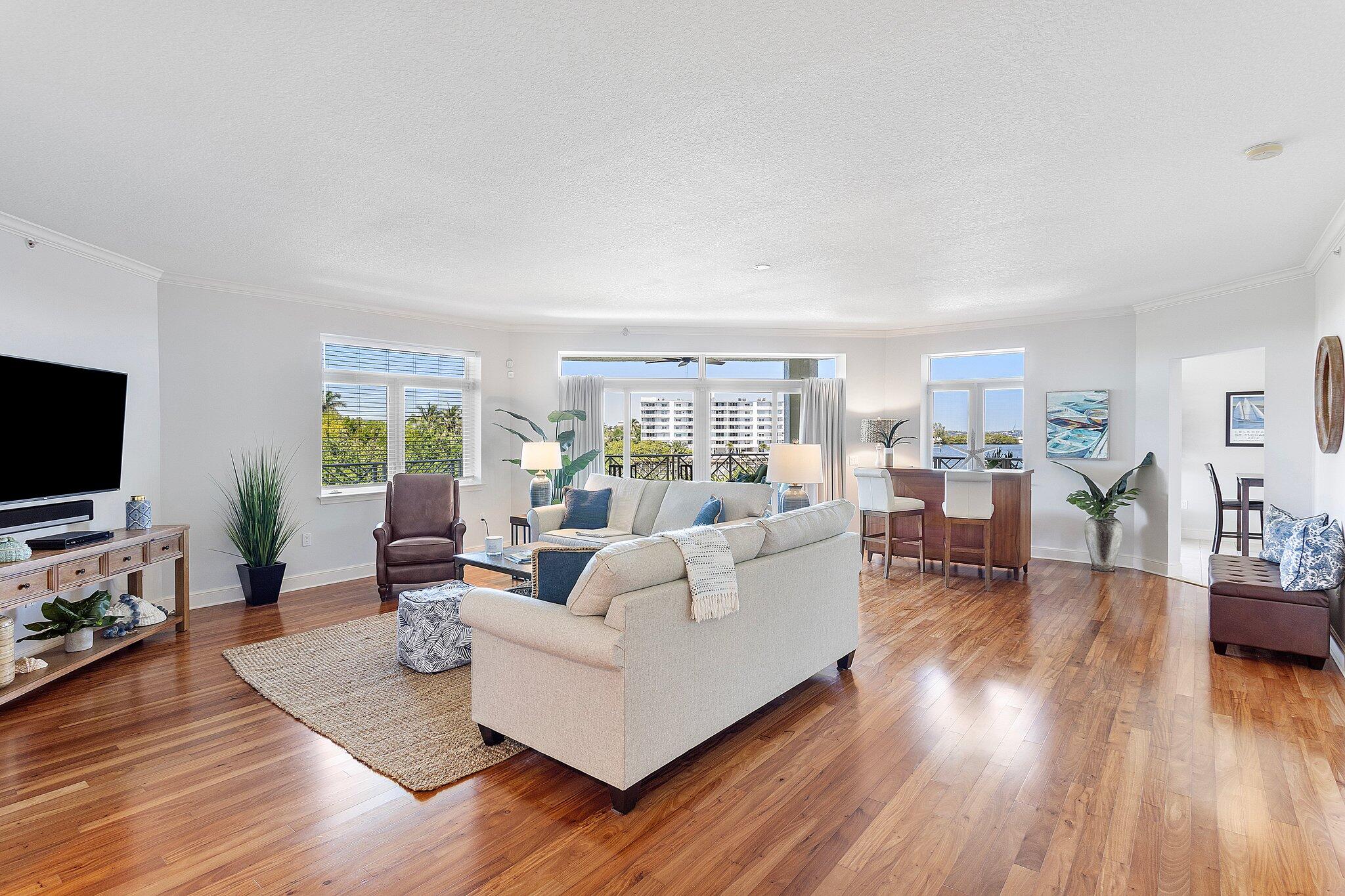 Bright living room with white sofas, hardwood floors, and windows overlooking water and nearby buildings.