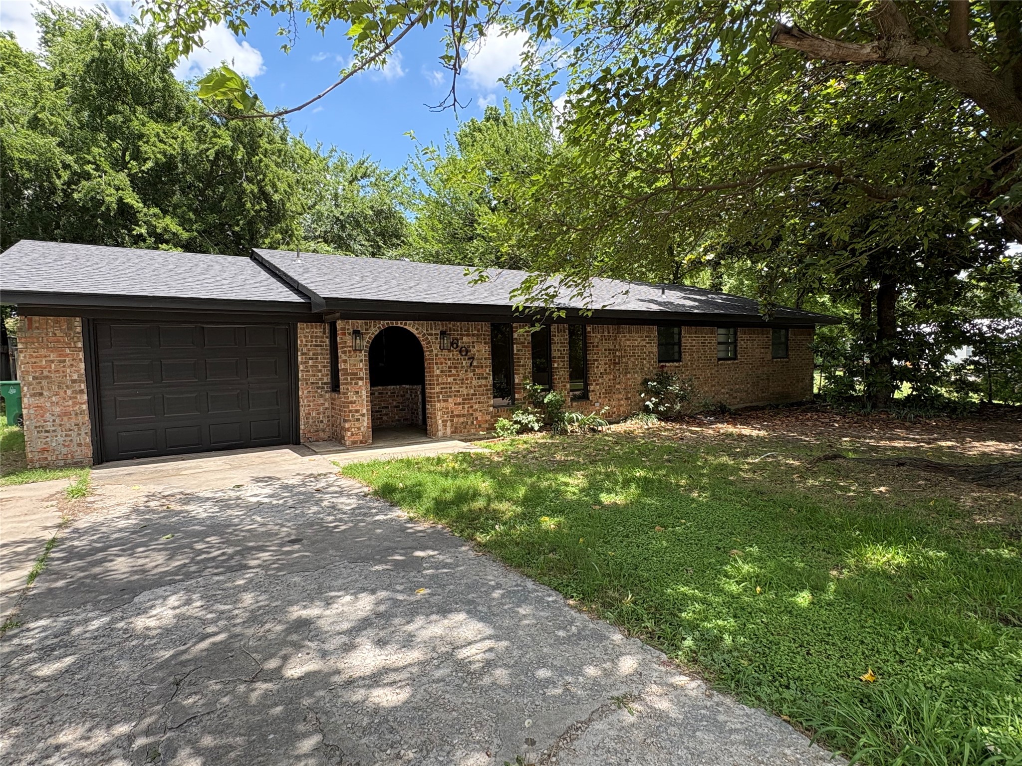Single-story brick house with a dark garage door and arched entry beneath mature trees.