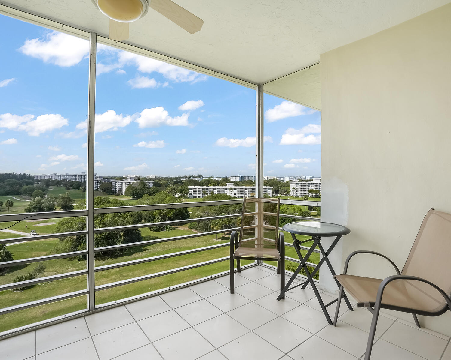 Glass-enclosed condo balcony with seating overlooking a golf course and residential buildings.