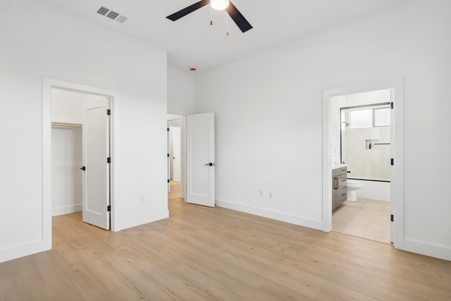 Empty bedroom with light oak floors, a black ceiling fan, and an attached bathroom.