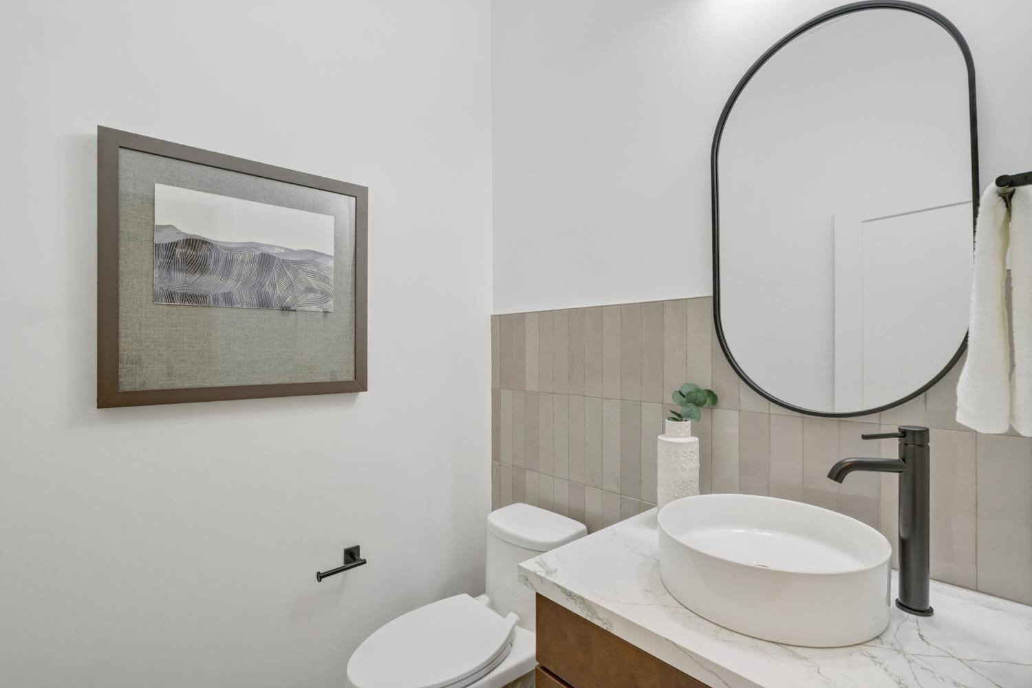 Bathroom vanity with a white vessel sink, dark faucet, tiled backsplash, and large oval mirror.