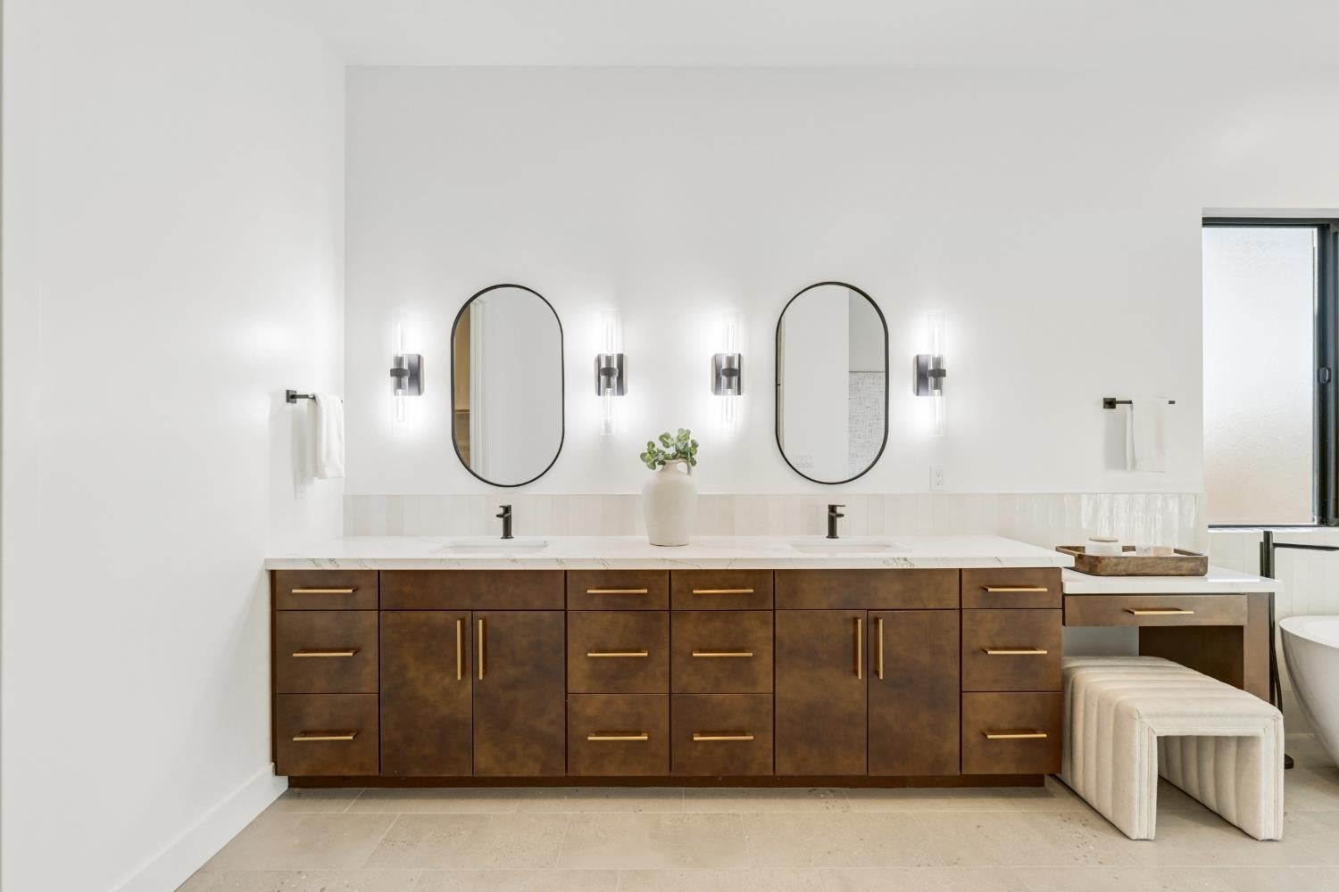 Modern bathroom with a dark wood double vanity, oval mirrors, and a cushioned stool.