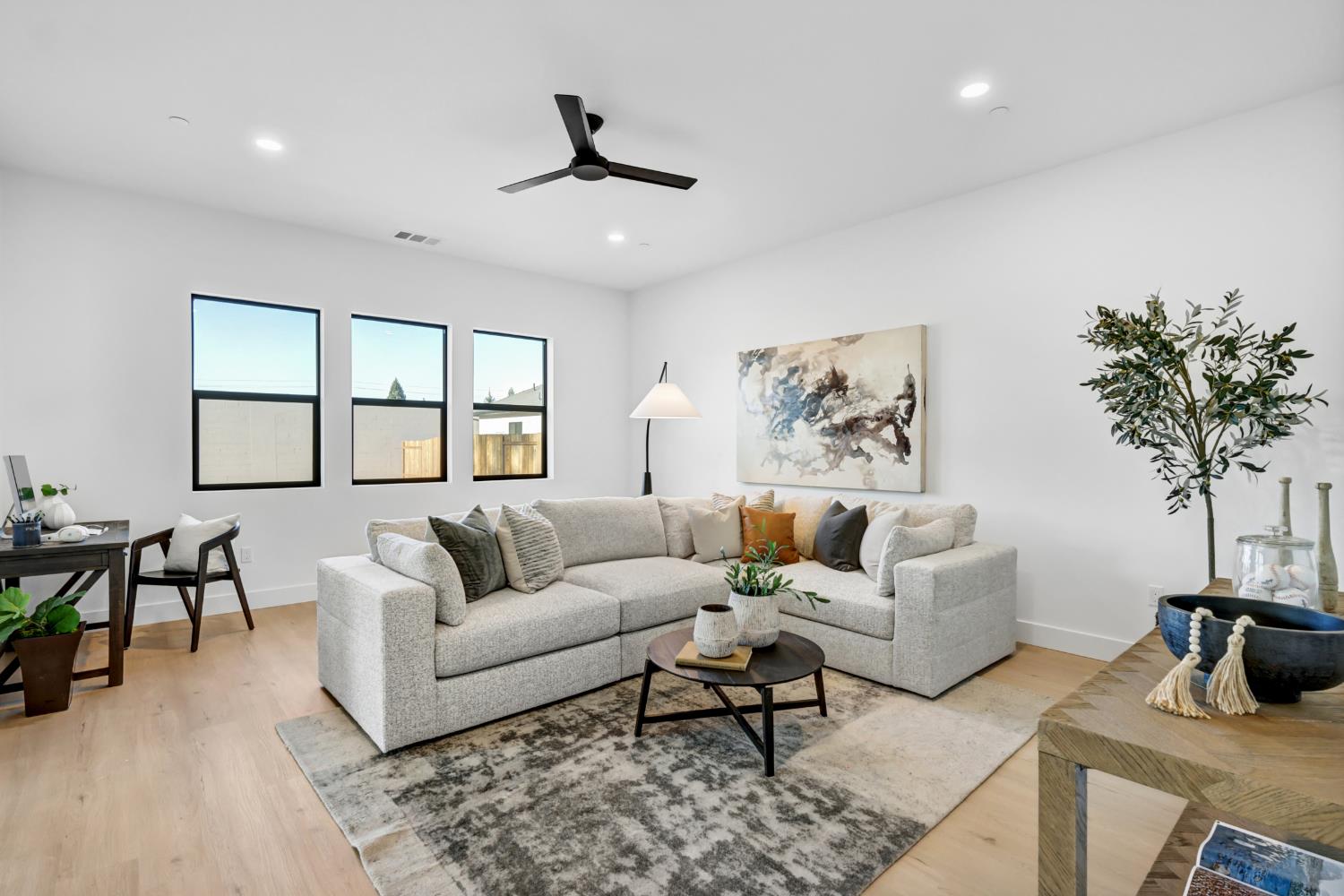 Staged living room with a light-gray sectional, abstract artwork, and three tall windows.
