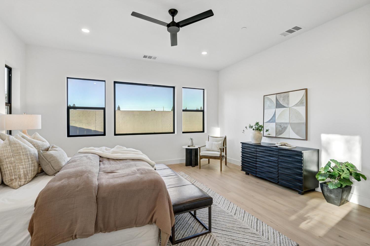 Bright staged bedroom with neutral bedding, three black-framed windows, and a dark dresser.