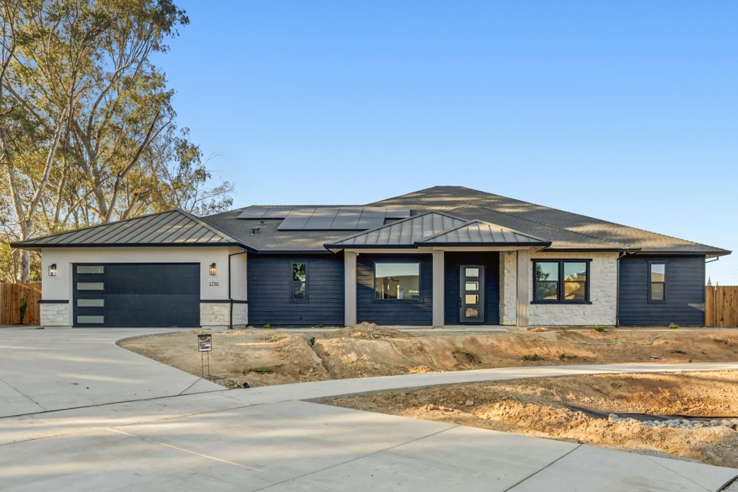 Single-story modern home with dark siding, stone accents, solar panels, and unfinished front landscaping.