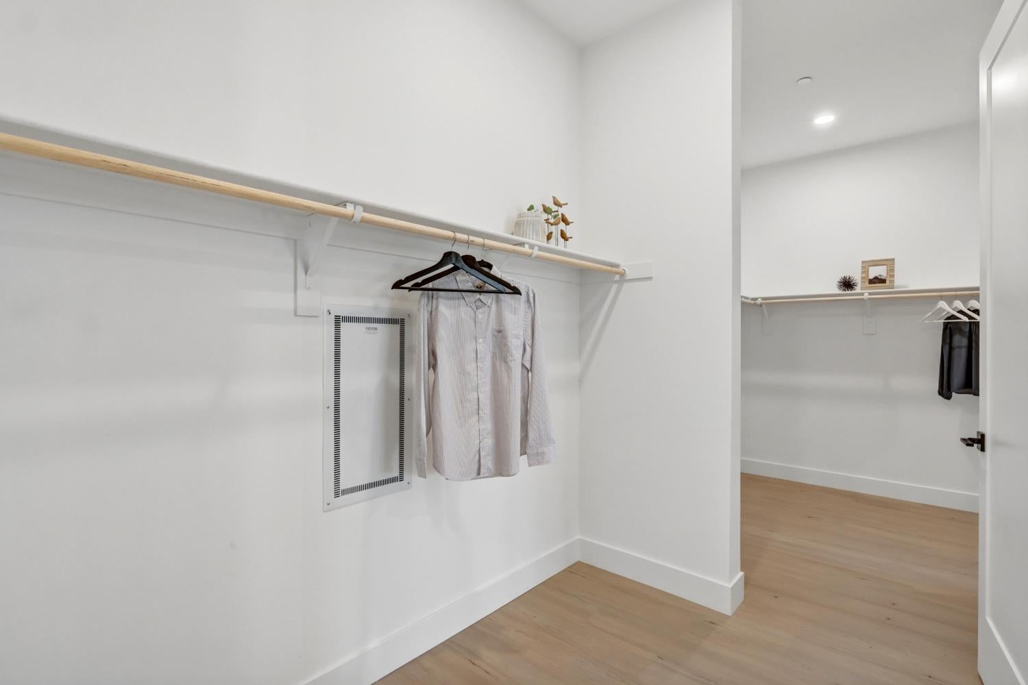 Walk-in closet with long hanging rods, a few garments, and light wood flooring.