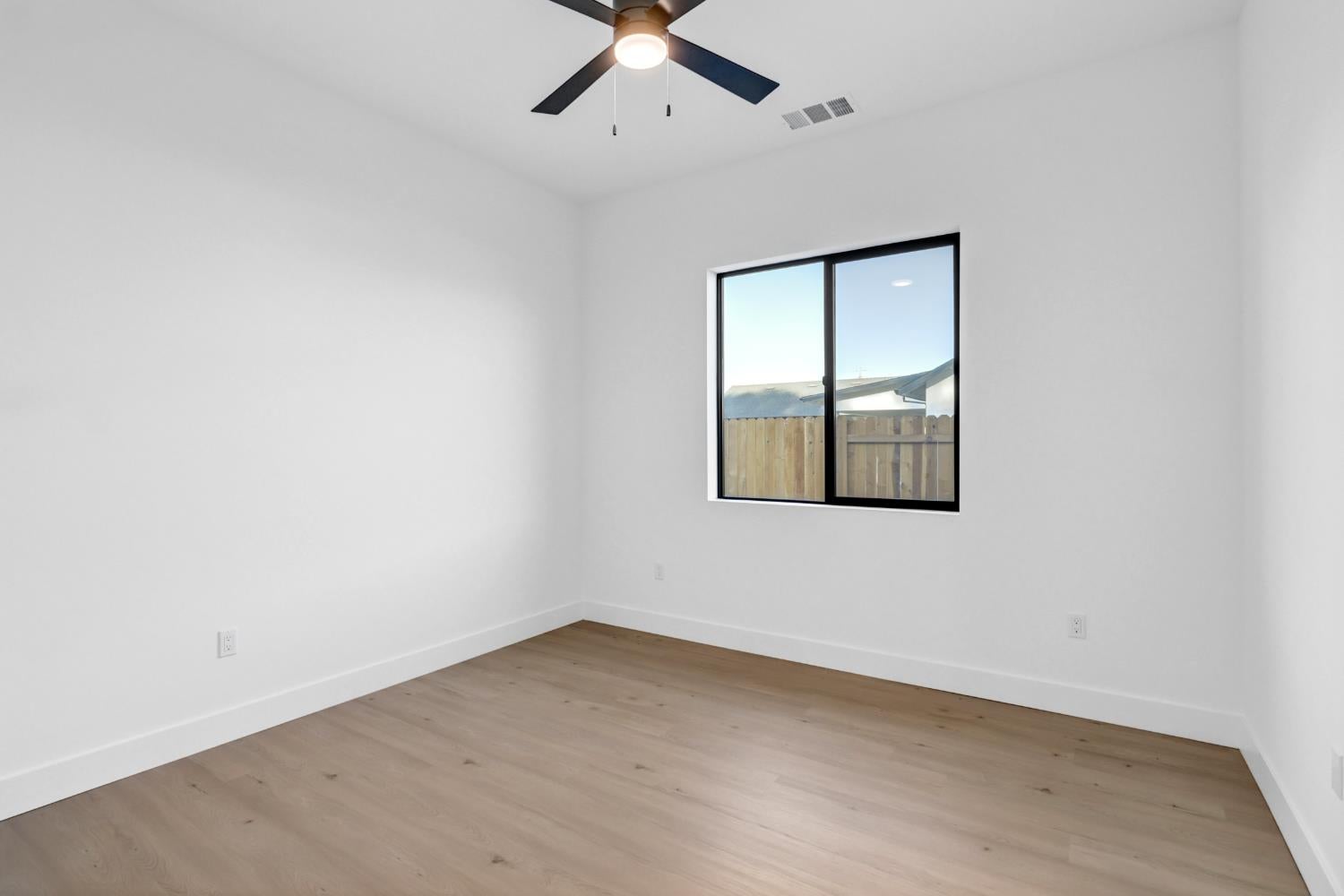 Empty room with light wood flooring, a black ceiling fan, and a window facing a fenced yard.