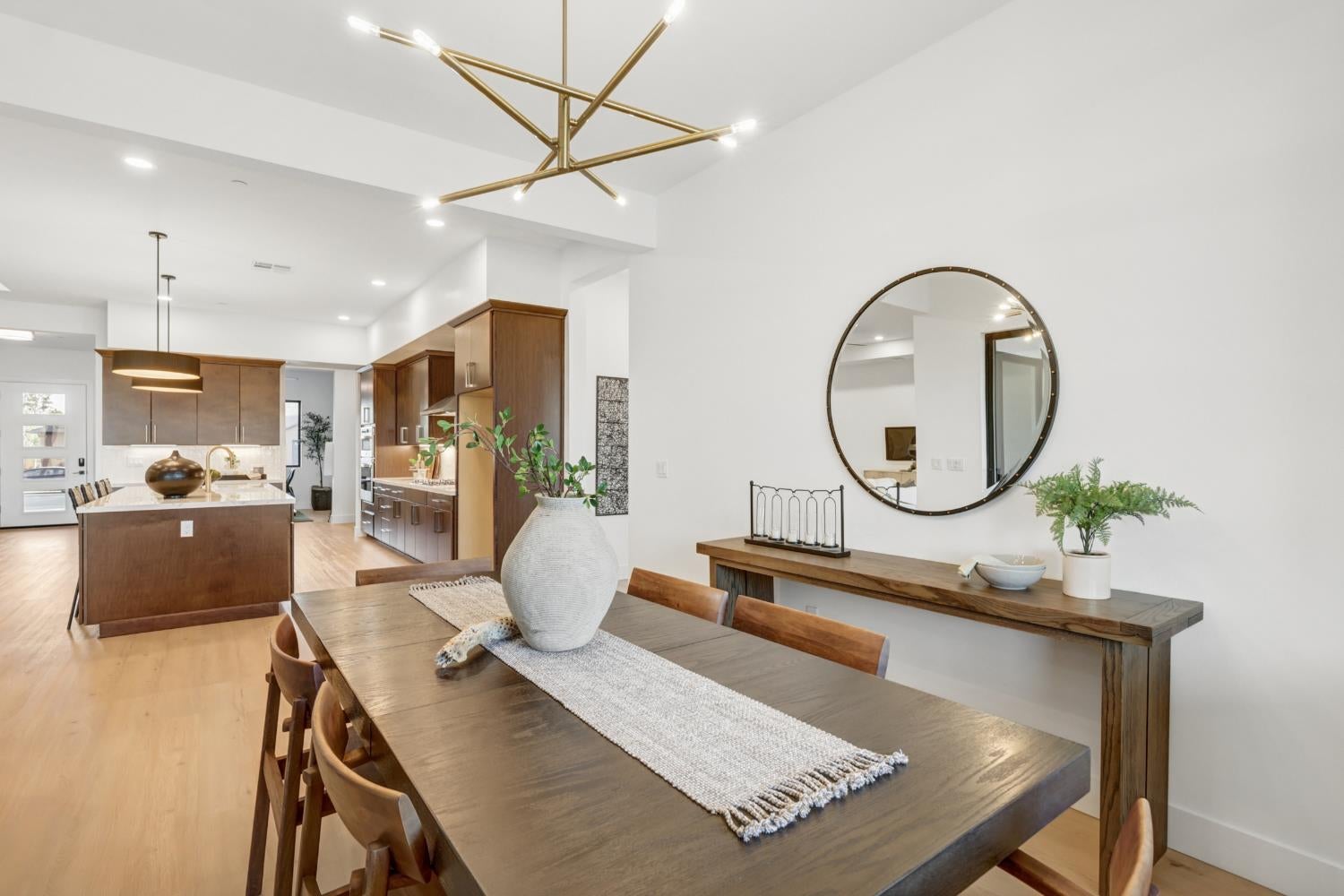 Open-plan dining room with a large wood table, round mirror, and kitchen with warm wood cabinetry.