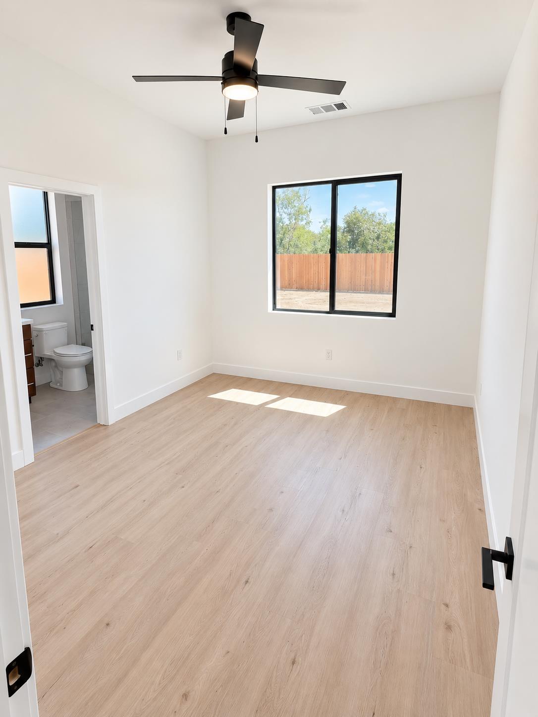 Empty bedroom with pale wood floors, a black ceiling fan, and a window overlooking a fenced yard.