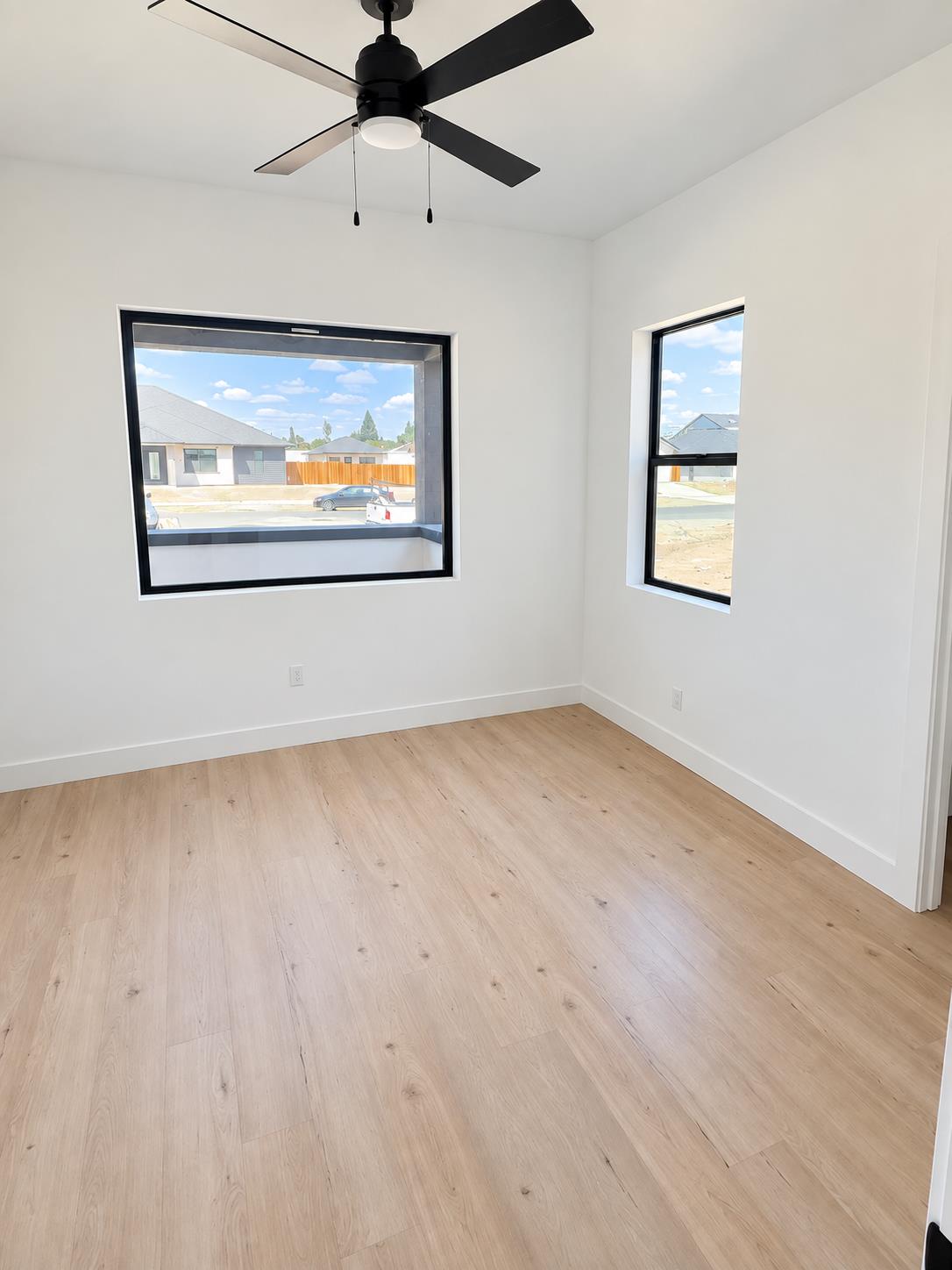 Empty room with pale wood floors, a black ceiling fan, and two windows overlooking neighboring homes.