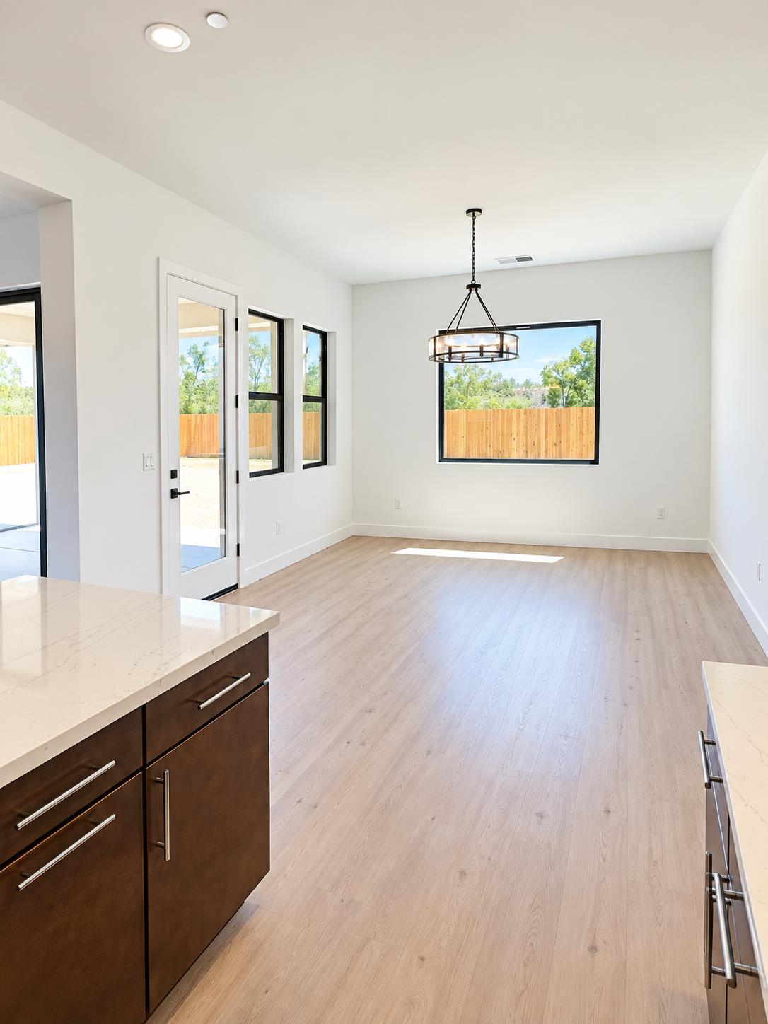 Vacant dining area with a ring chandelier, light wood floors, and windows overlooking a fenced yard.