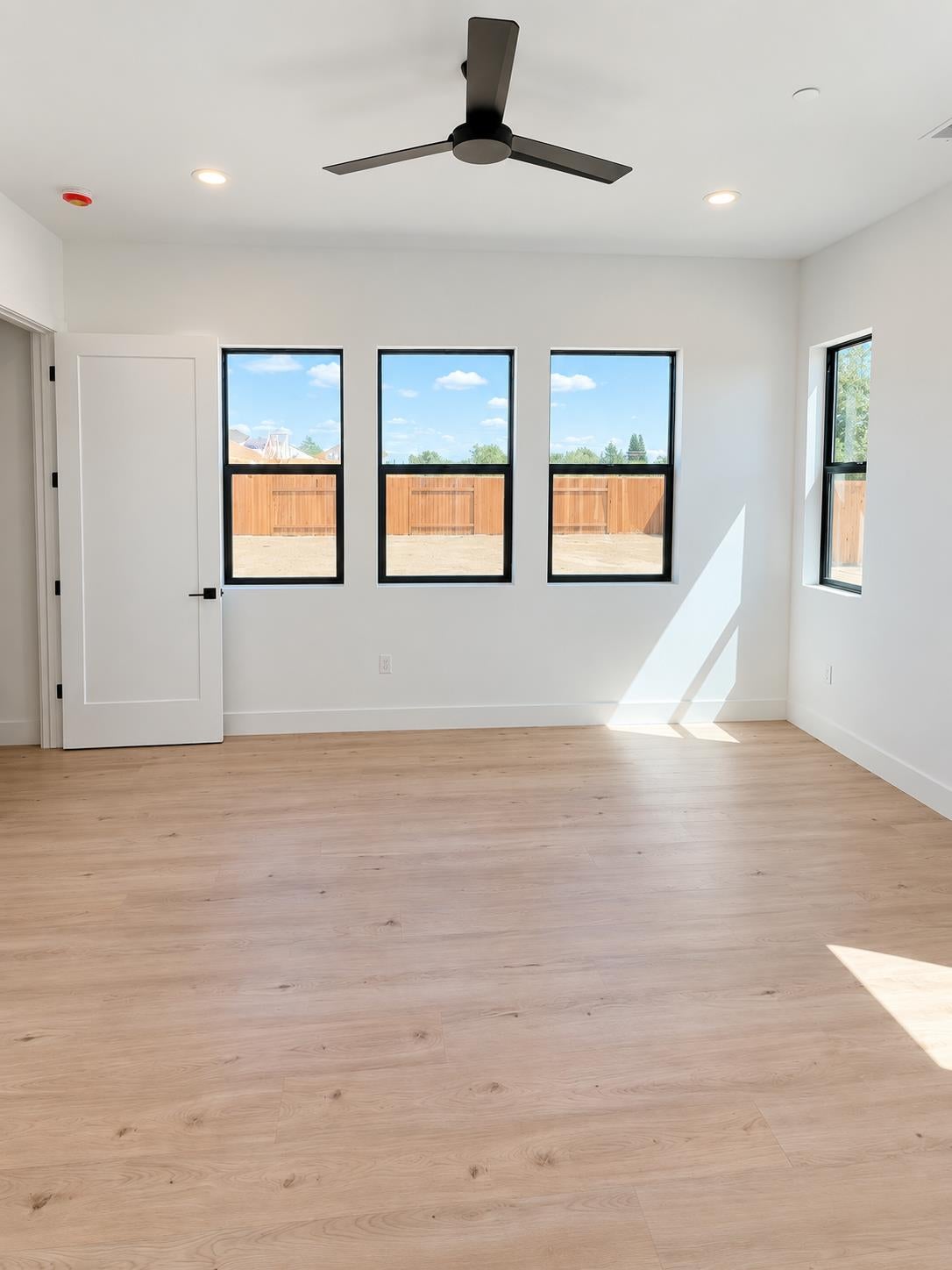 Empty room with light wood floors, a black ceiling fan, and tall windows overlooking a fenced yard.