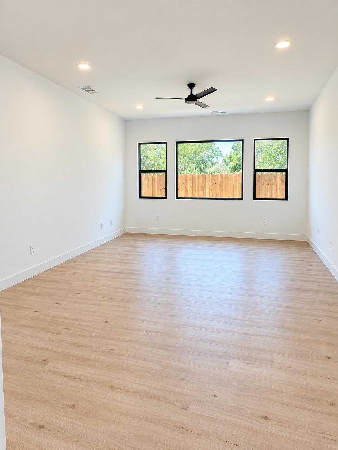 Empty room with light plank flooring, black ceiling fan, and three windows facing a fenced yard.
