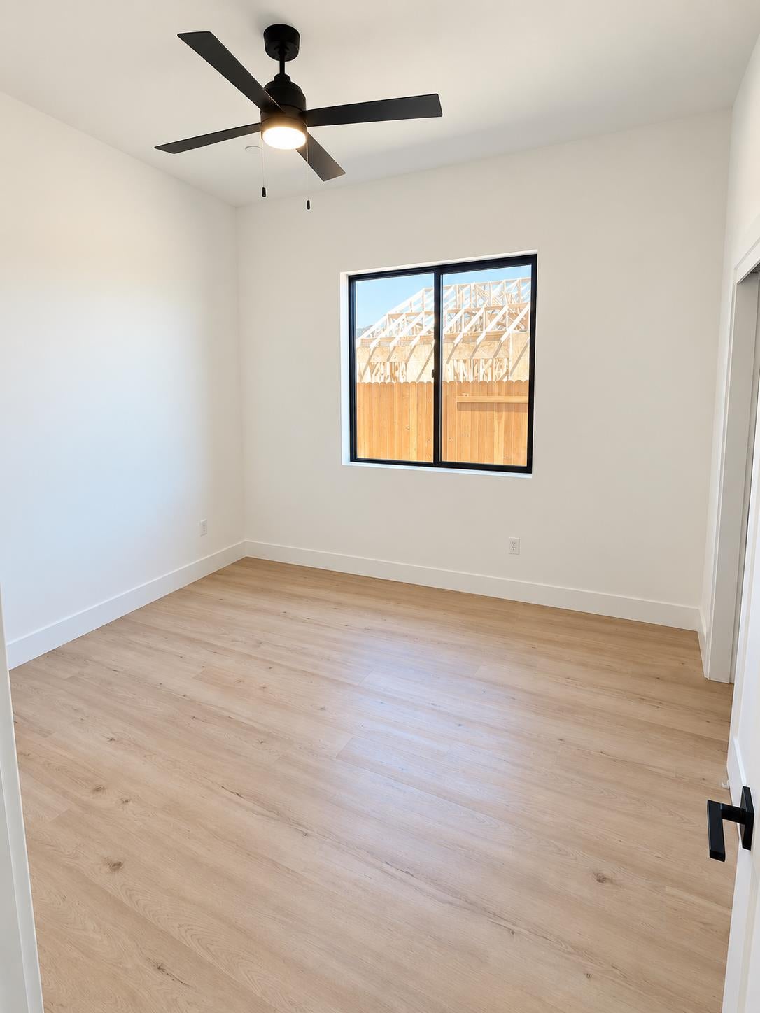 Vacant room with pale wood floors, a black ceiling fan, and a window overlooking a wooden fence.