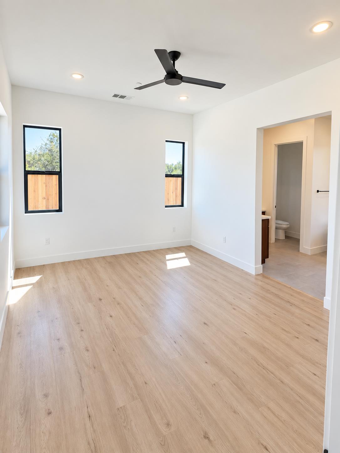 Empty bedroom with light wood floors, two narrow windows, and an open doorway to an en-suite bathroom.