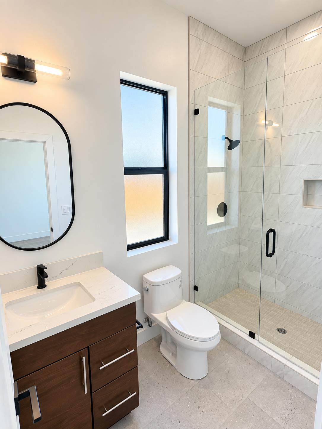 Modern bathroom with a marble-look glass shower, dark wood vanity, and oval mirror.