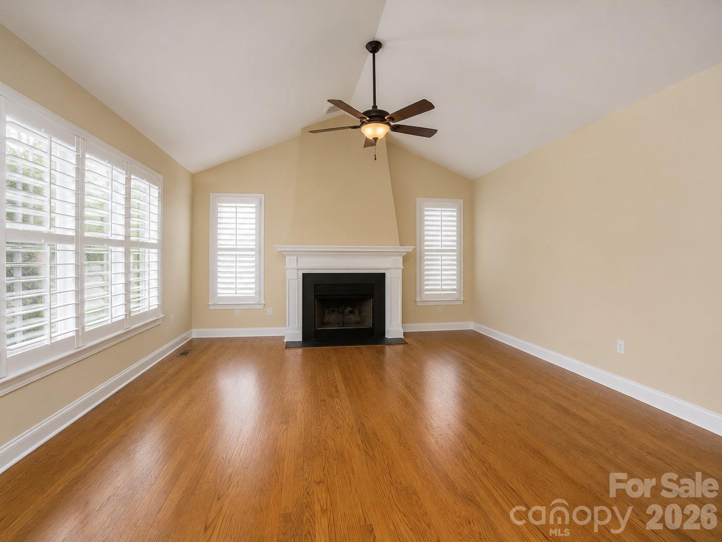 Empty living room with hardwood floors, plantation shutters, a white fireplace, and a ceiling fan.