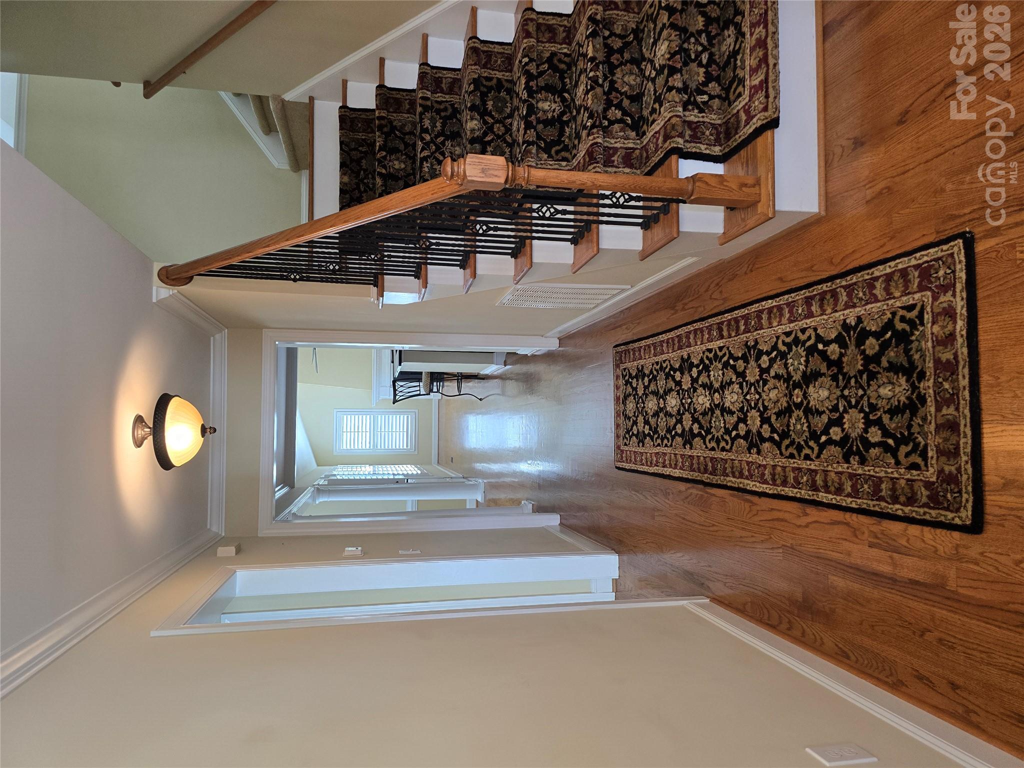 Foyer hallway with hardwood floors, a patterned runner, and a carpeted staircase with black iron balusters.