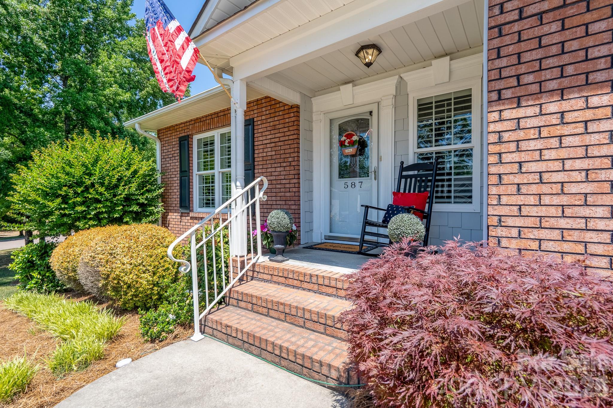 Brick home entrance with covered porch, rocking chair, American flag, and landscaped steps.