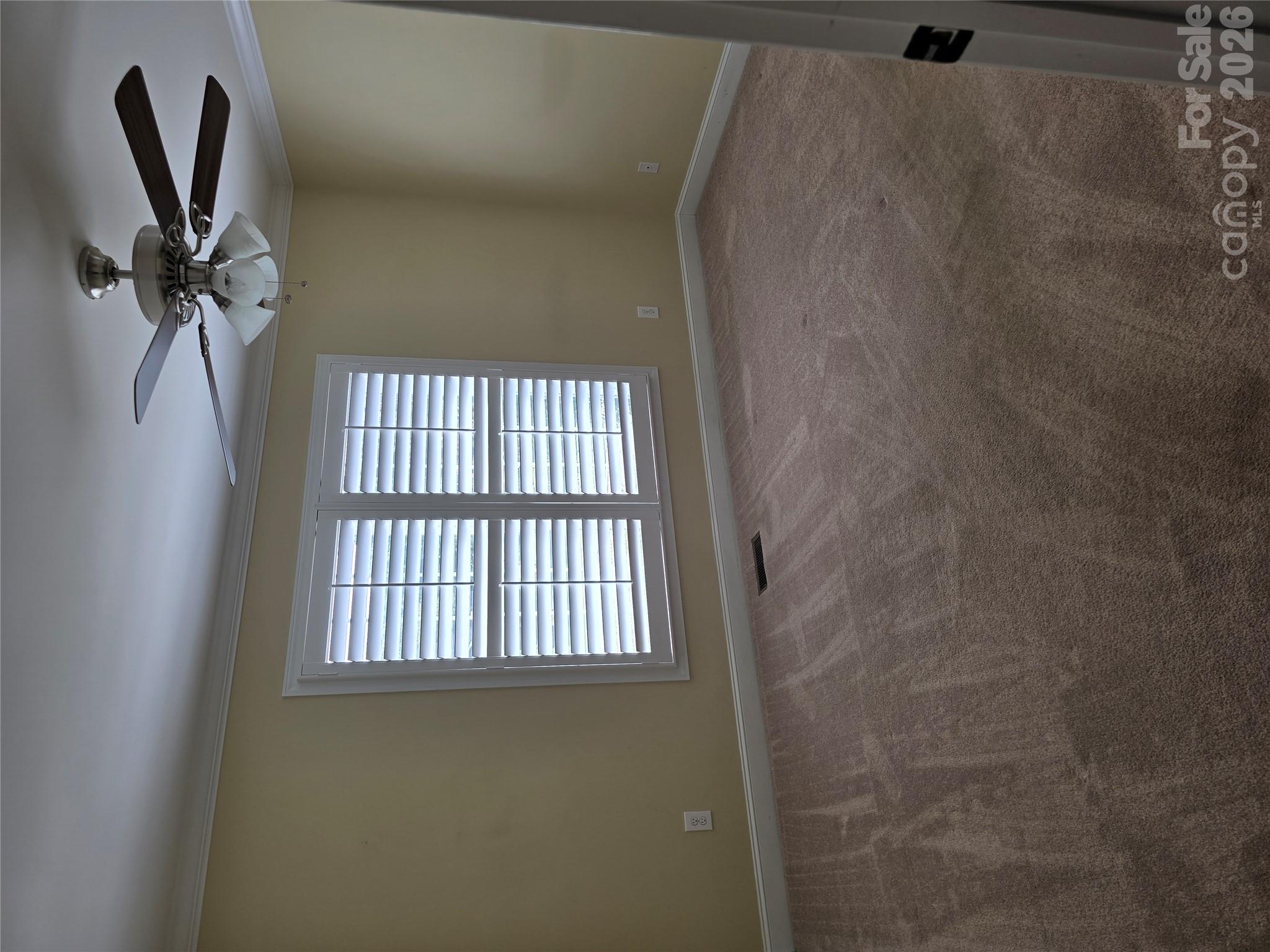 Empty carpeted room with plantation-shuttered window and ceiling fan with lights.