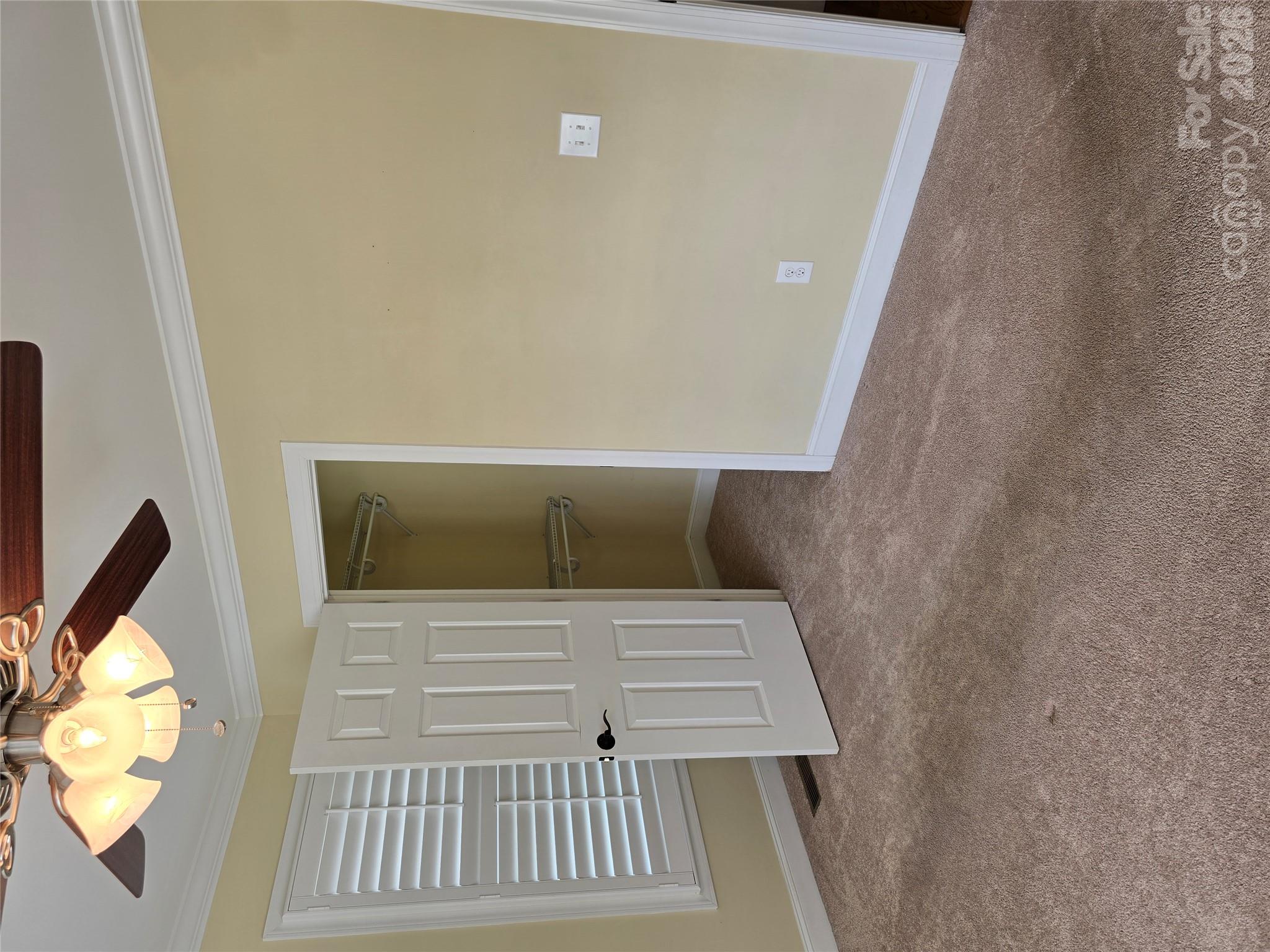 Empty bedroom with beige carpet, a lit ceiling fan, shuttered window, and open closet.