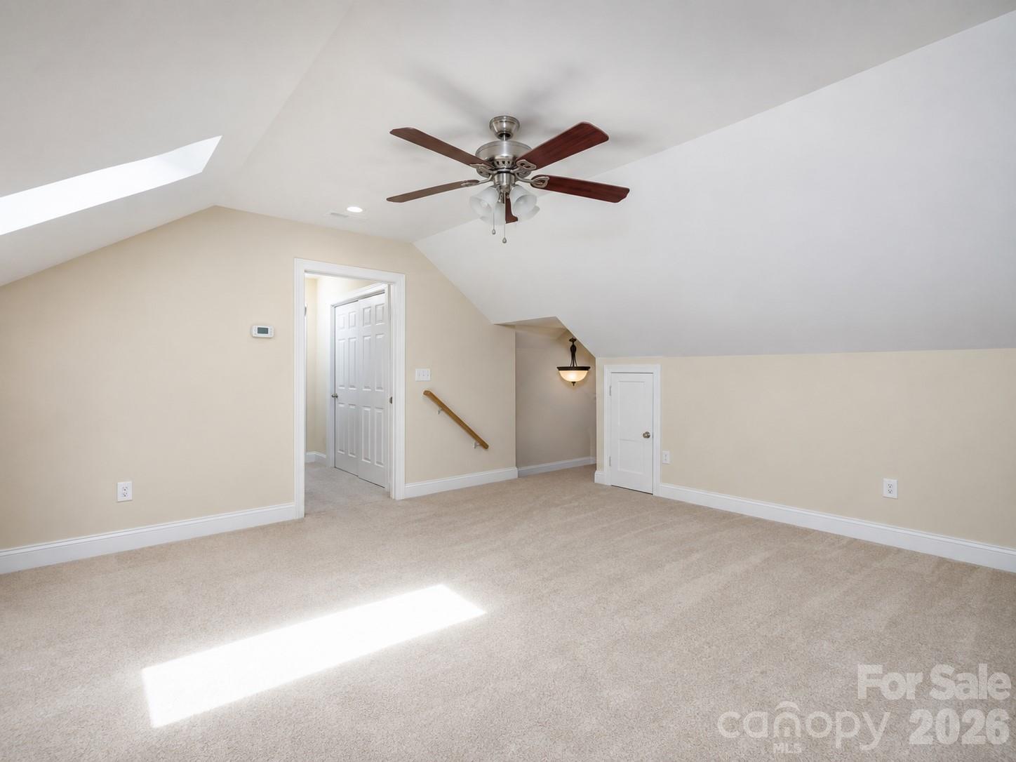 Empty carpeted upstairs room with a sloped ceiling, ceiling fan, and sunlight from a skylight.