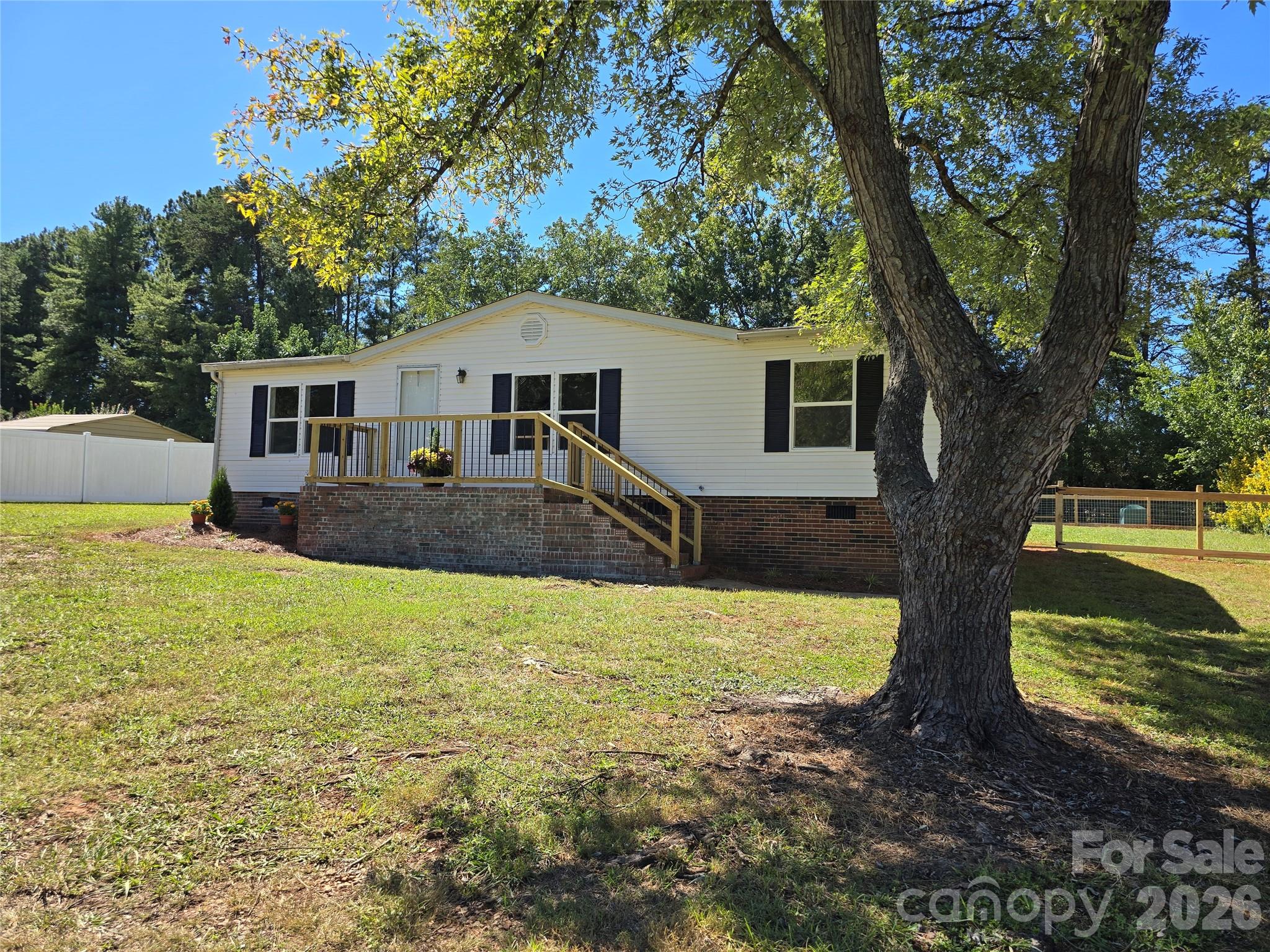 White single-story home with a raised porch, brick foundation, and large shade tree in the front yard.