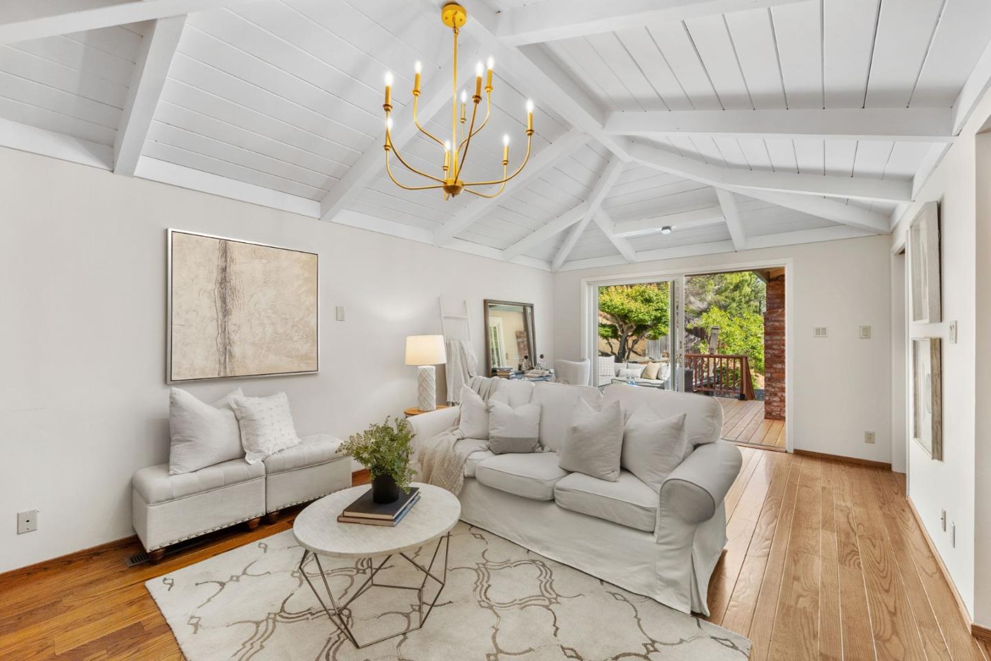 Living room with a vaulted beam ceiling, white sofas, and sliding doors opening to a leafy deck.