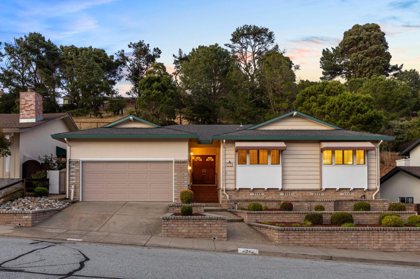 Single-story home with a double garage, warmly lit entry, and brick front planters at dusk.