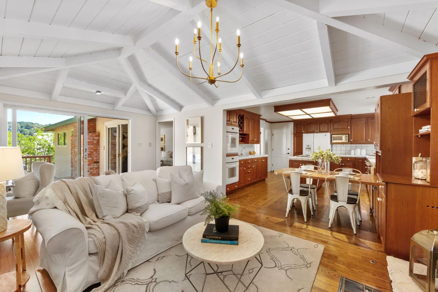 Open-plan living room and kitchen with a vaulted beamed ceiling, brass chandelier, and warm wood cabinetry.