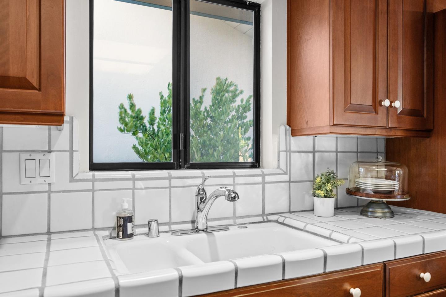Kitchen sink beneath a window, framed by white tile counters and warm wood cabinets.
